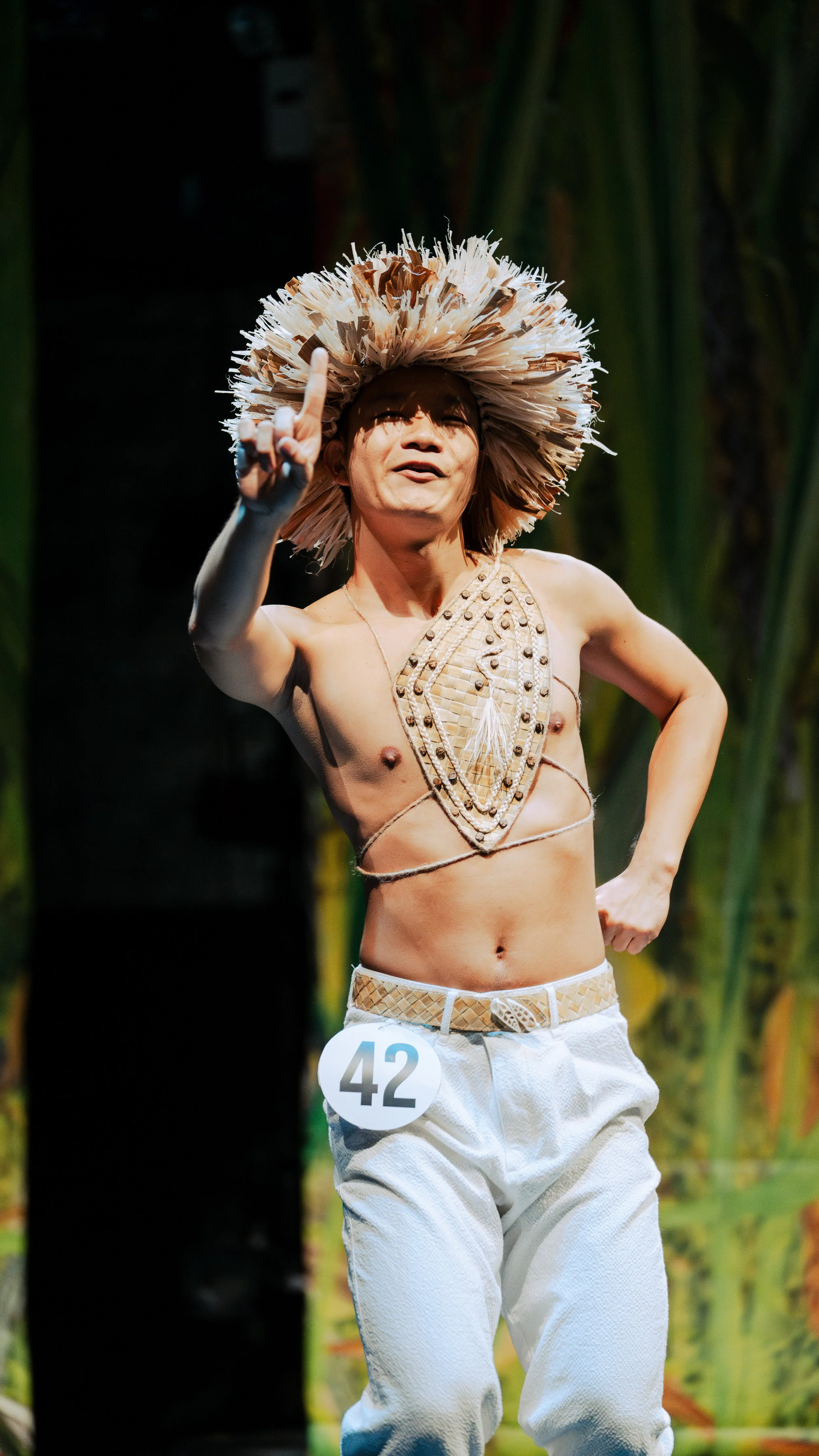 Person performing on stage wearing traditional attire with a numbered badge, straw headdress, and decorative chest piece.