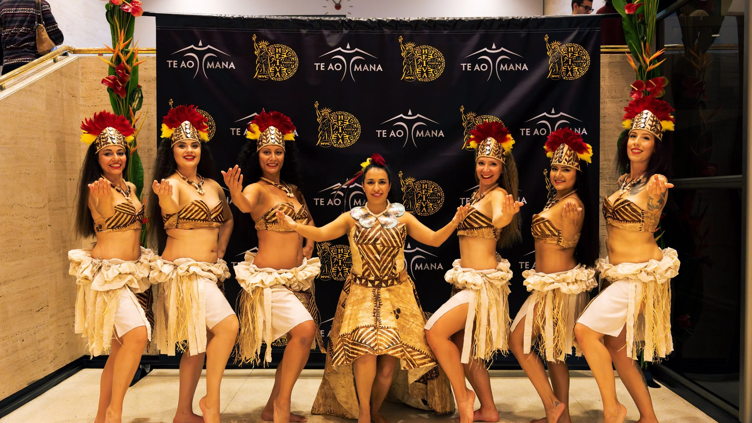 Group of women in traditional Polynesian dance costumes posing in front of a branded backdrop.