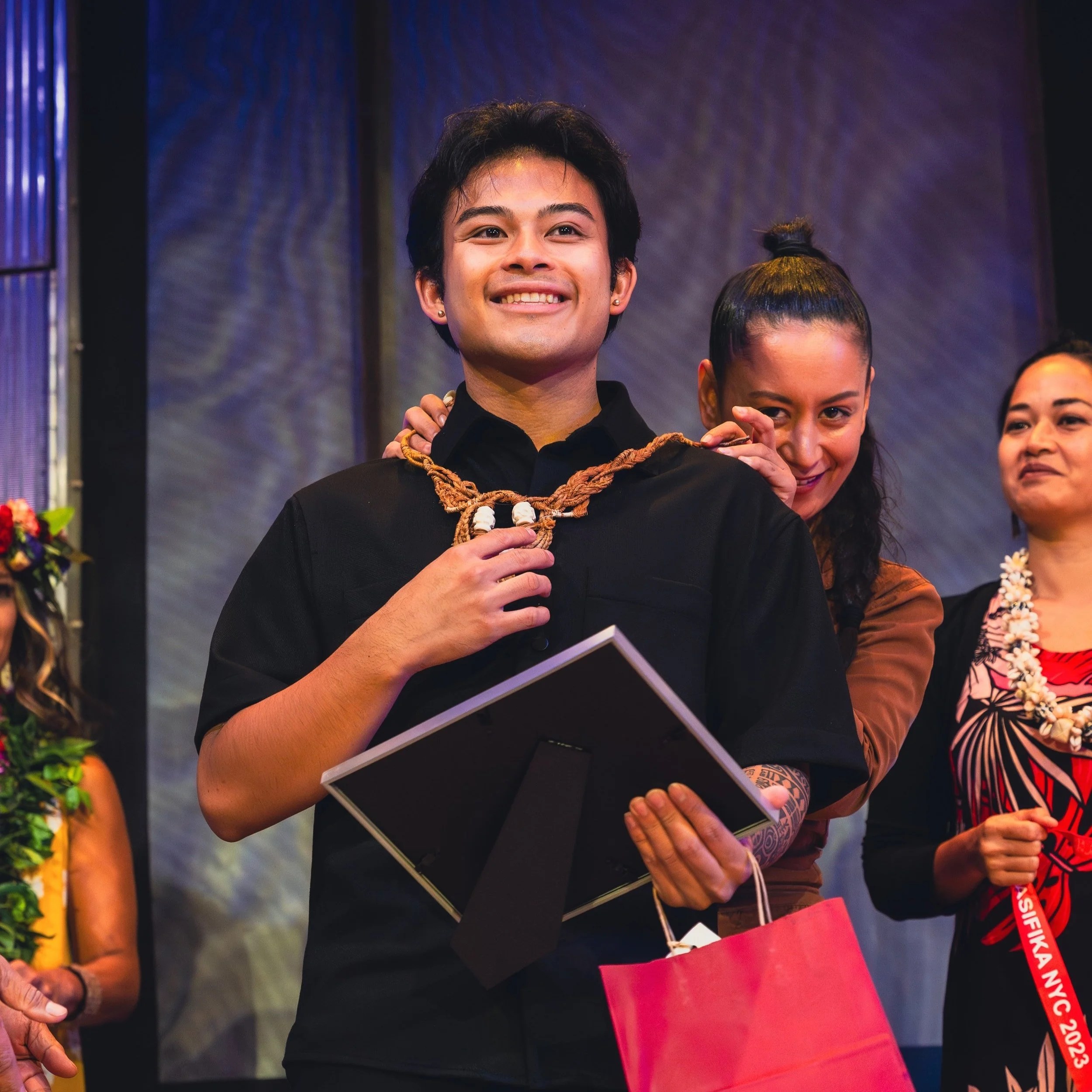Group on stage celebrating, one person receiving an award with a wooden necklace and smile, holding a red gift bag and framed item.