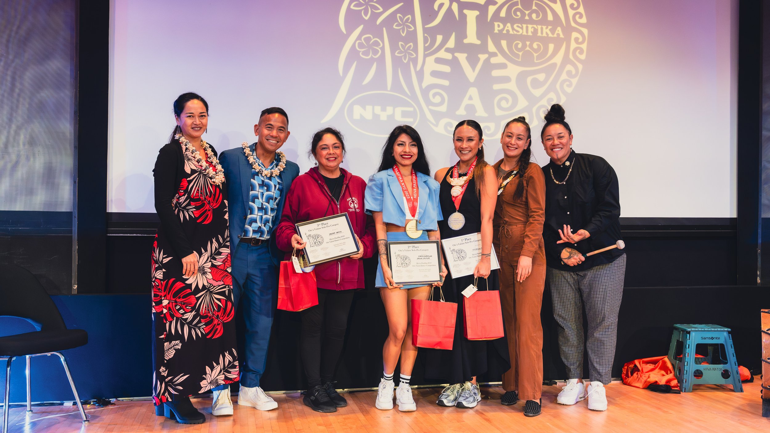 Group of seven people posing with awards and leis, standing on a stage with a Pasifika emblem in the background.
