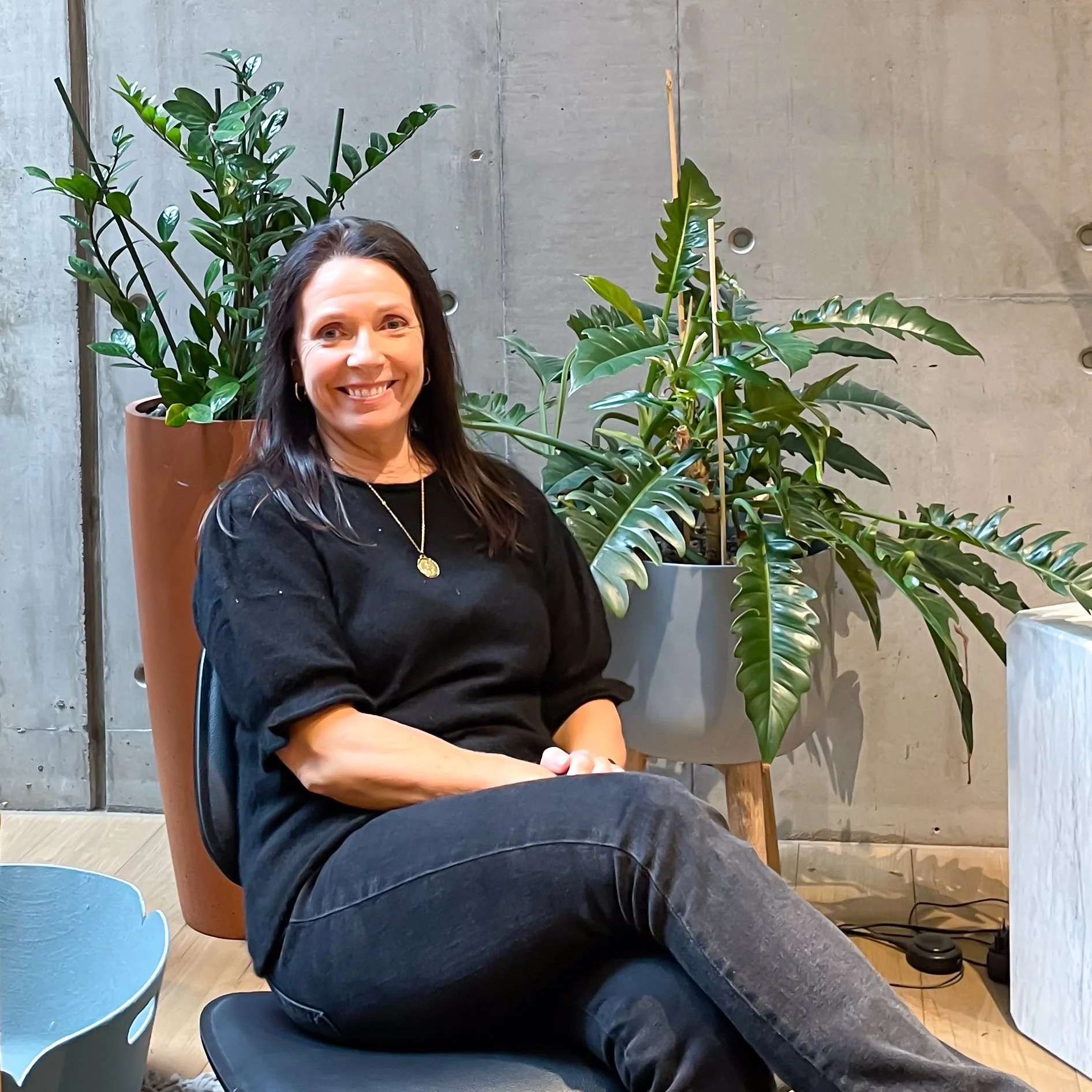 En kvinne sitter i en stol foran en vegg med to store grønne planter i potter.