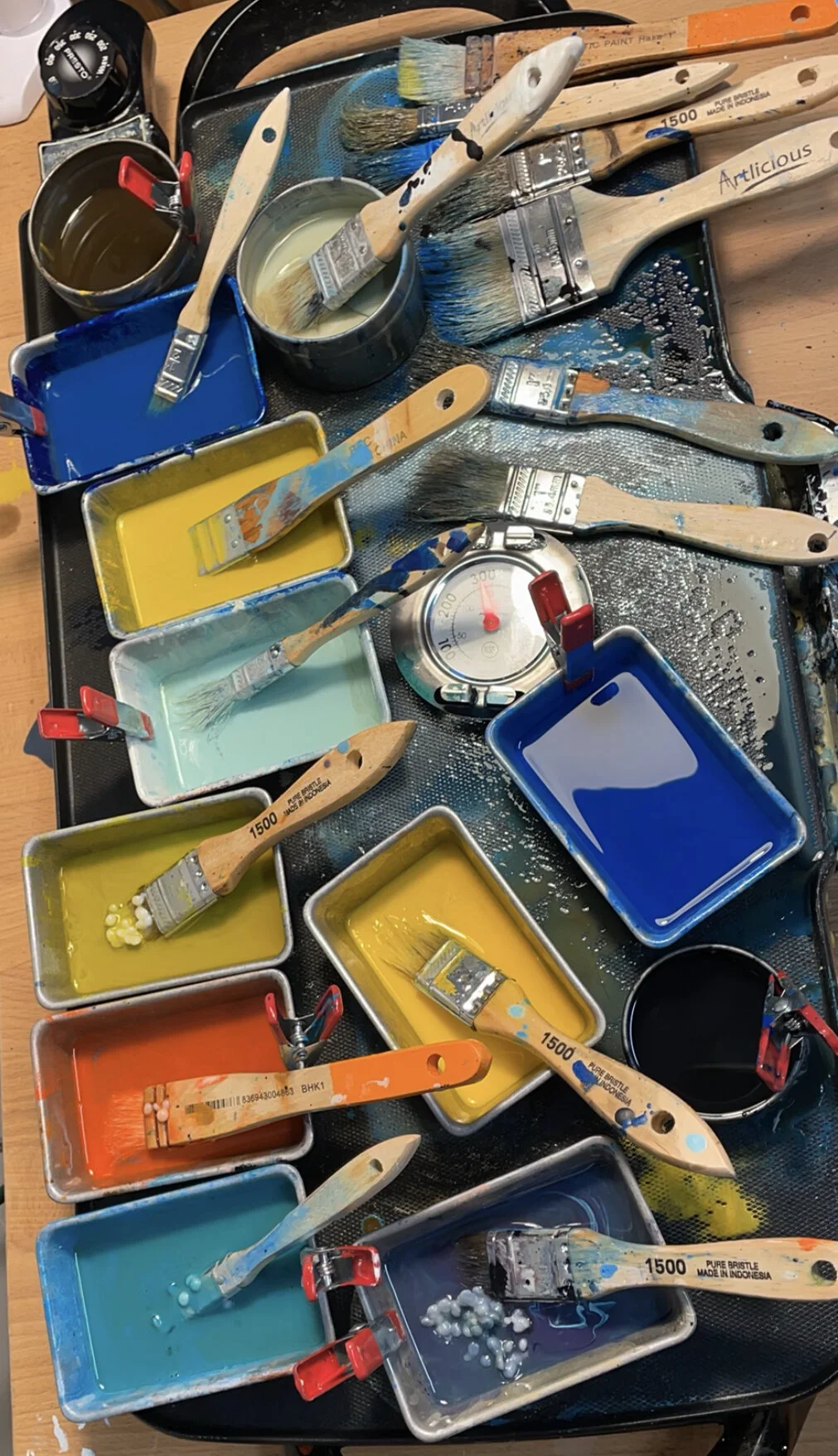A painting palette with several paintbrushes dipped in various colors of paint, some in small containers, with a thermometer nearby, on a black surface.