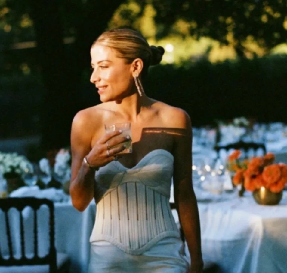 Woman in elegant strapless dress holding a glass, outdoors at a formal event with decorated tables and floral centerpieces