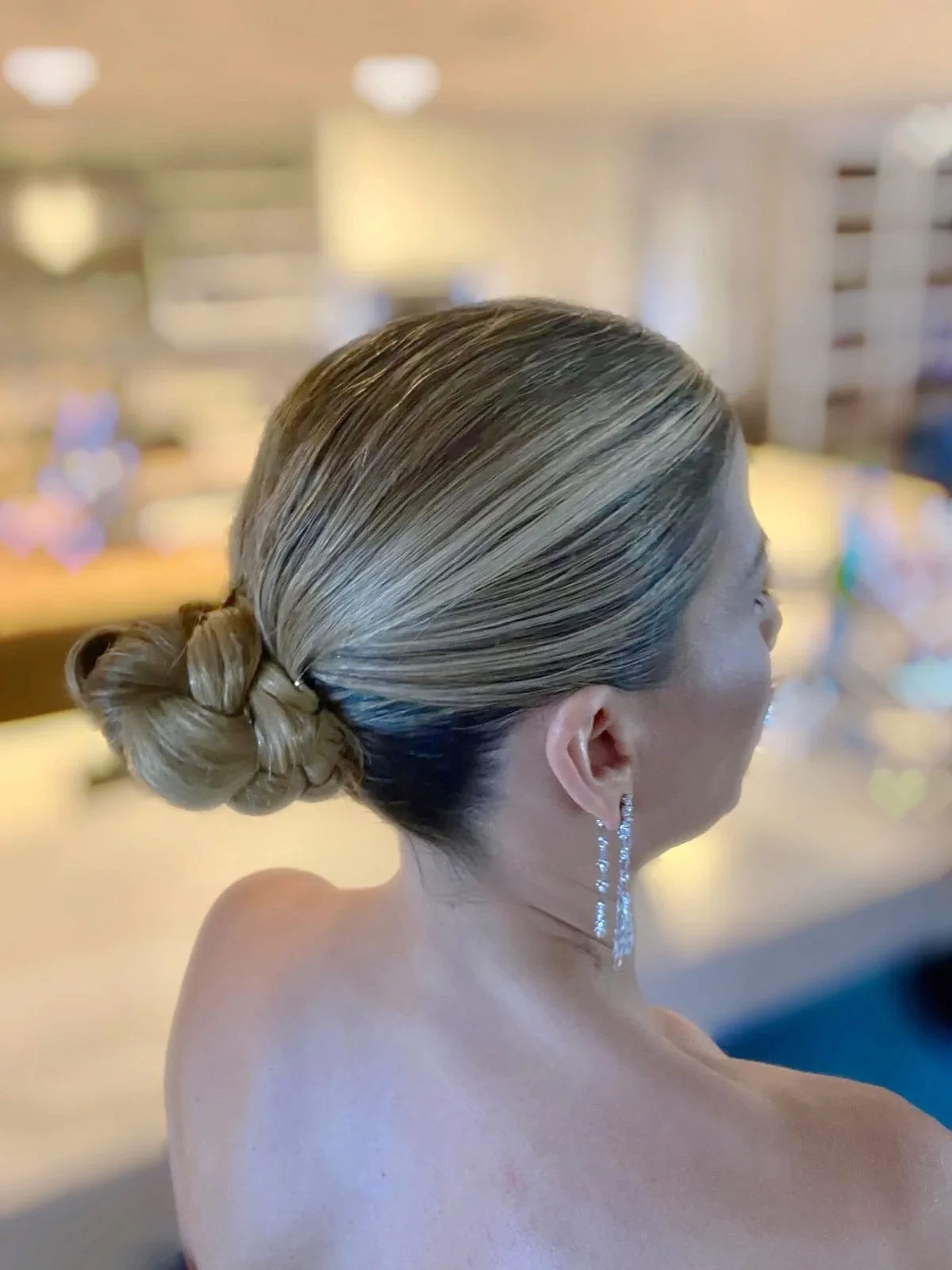 A woman with blonde hair styled in a neat bun, wearing long, dangling diamond earrings, in a softly lit indoor setting.