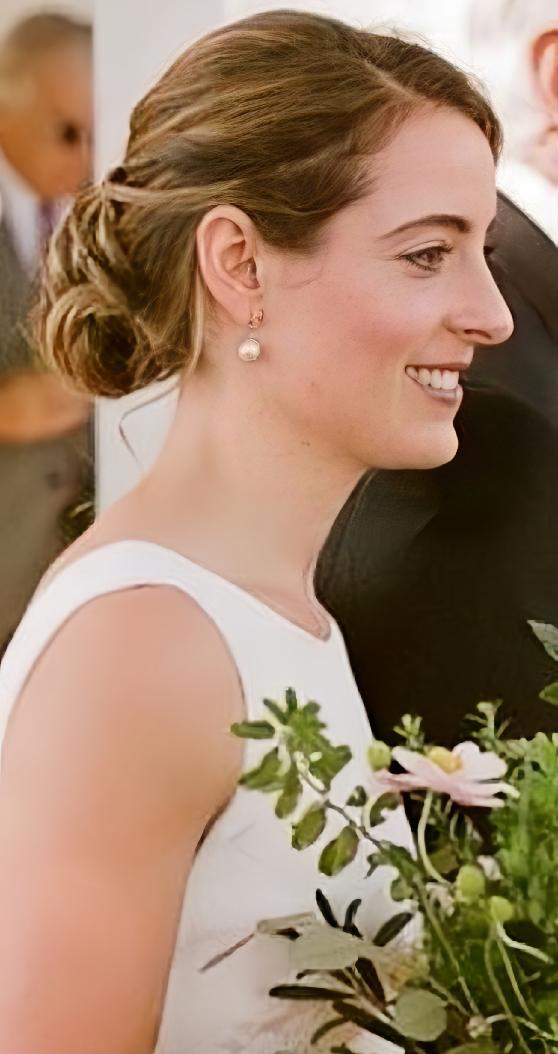 A woman with light brown hair styled in an updo, smiling and wearing pearl earrings, holding a bouquet of green foliage and pink flowers, dressed in a white sleeveless dress, at a formal event or wedding.
