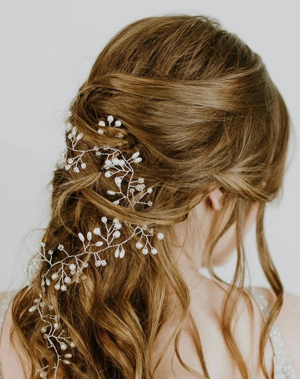 A woman with long, wavy, light brown hair styled in loose waves, decorated with a white floral and pearl hair accessory, with a few strands framing her face.