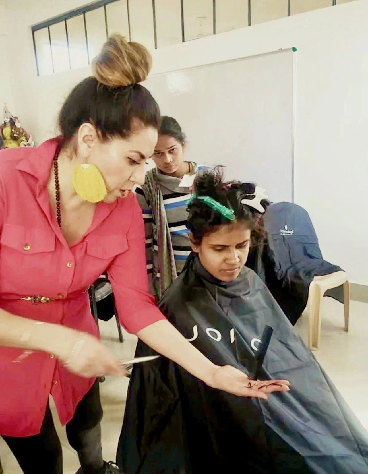 A woman giving a haircut to a young girl with hair clips in her hair, while a girl in the background watches.