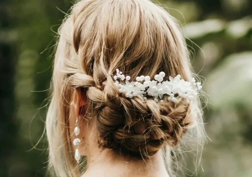 Back view of a woman with an intricate braided updo hairstyle decorated with white floral hair accessories and long pearl earrings, outdoors with greenery in the background.