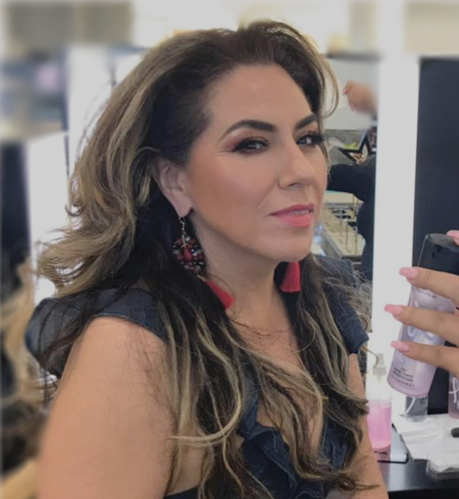 A woman with long wavy hair and earrings taking a selfie in a salon or makeup studio.