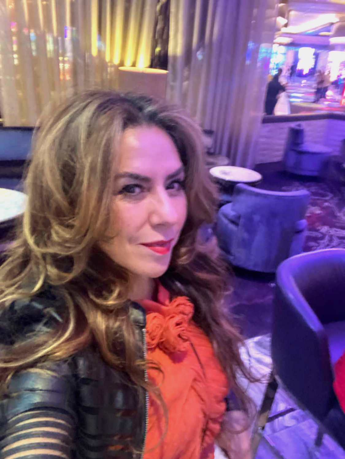 A woman with long, wavy hair and red lipstick taking a selfie inside a dimly lit, colorful lounge or casino with purple, yellow, and blue lighting.
