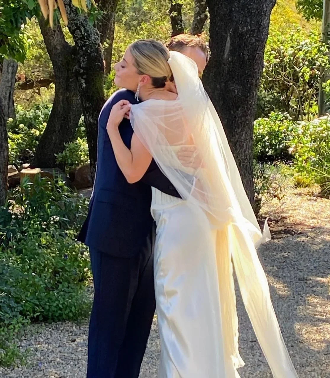 A bride and groom embrace outdoors among trees, with the bride wearing a white wedding gown and a chignon de marine (bridal bun)  with a veil  during daytime in Napa Valley.