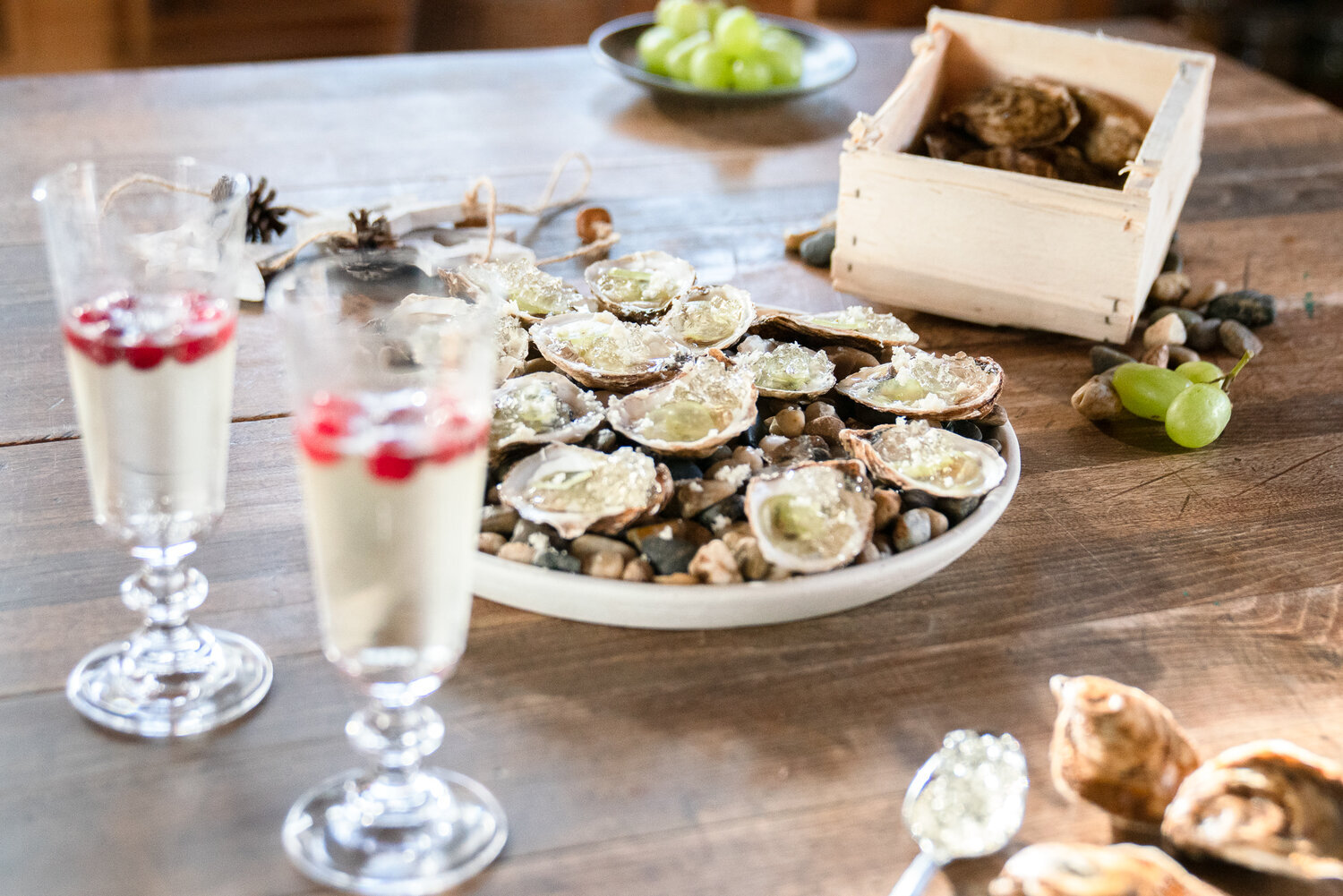 Une table en bois avec des huîtres dans une assiette, deux verres de boisson avec des baies rouges, et une boîte en bois contenant des coquilles vides. Des raisins verts sont visibles à l'arrière-plan.