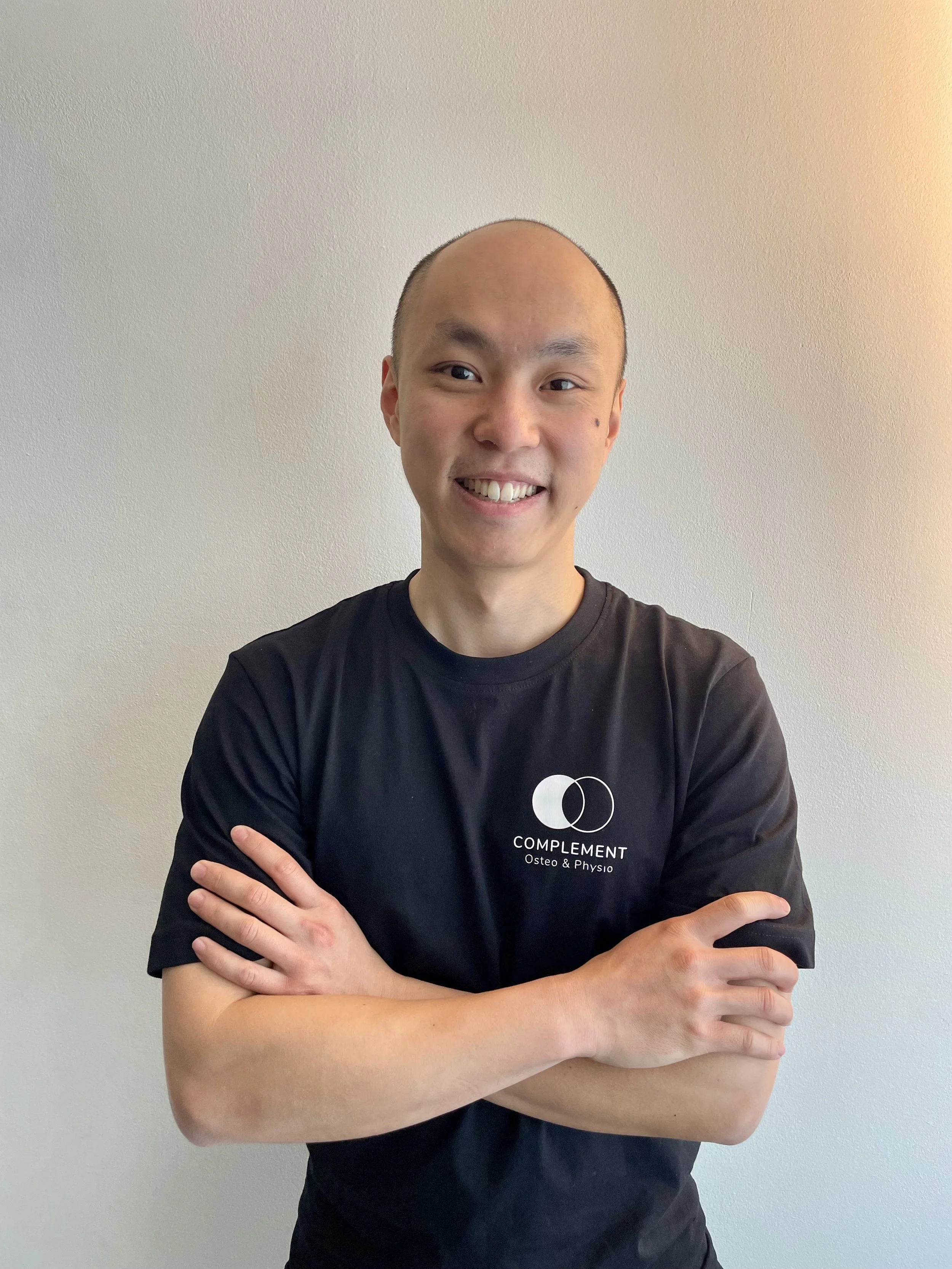 A smiling person with a shaved head wearing a black T-shirt that says 'COMPLEMENT Osteo & Physio', standing with crossed arms in front of a plain wall.