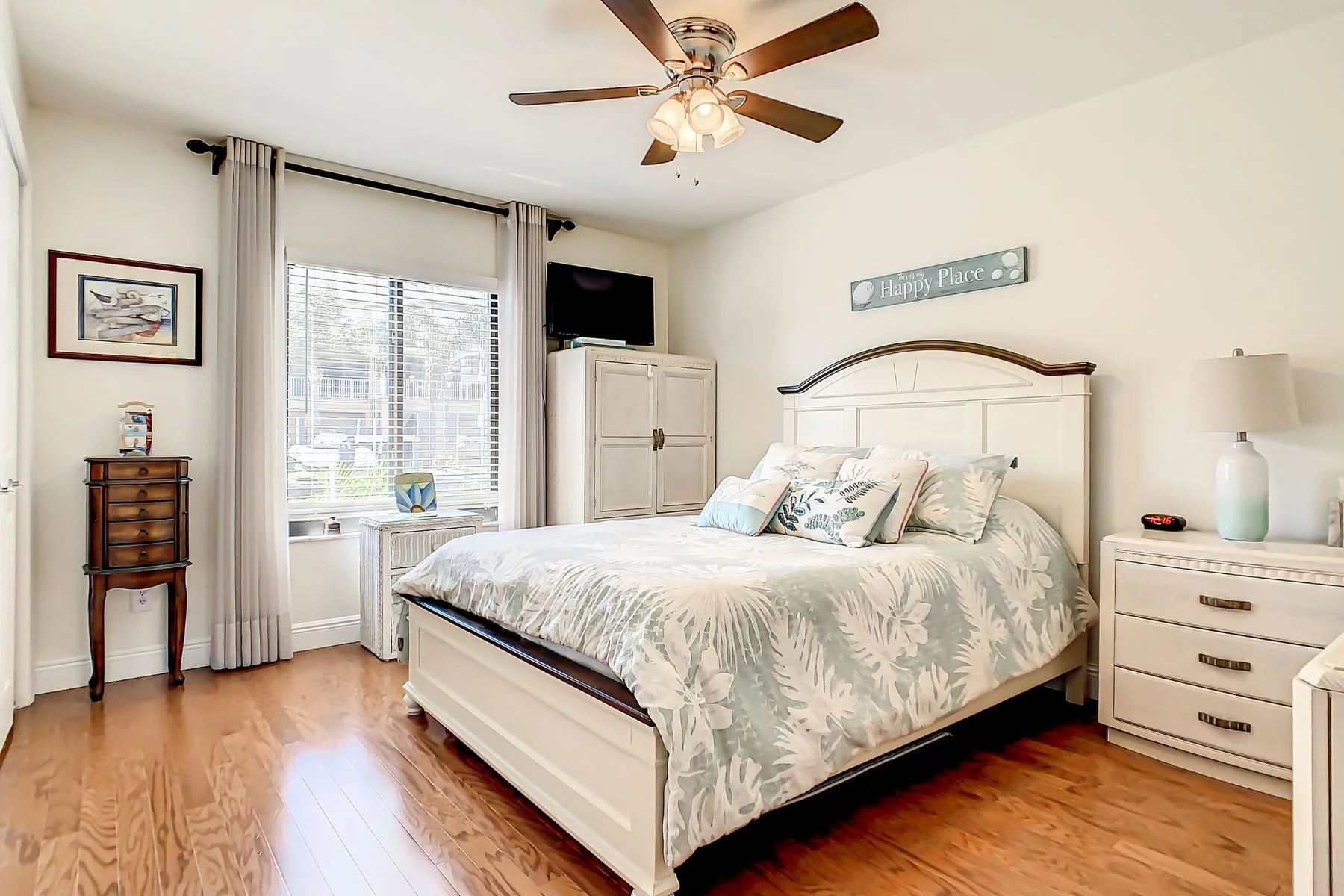 A bedroom with a white bed, floral bedding, and multiple pillows. There is a white nightstand with a lamp and digital clock, a white dresser, and a small wooden side table. A large window with blinds and curtains lets in natural light, and a ceiling fan with lights is above. A sign reads 'Happy Place' on the wall.