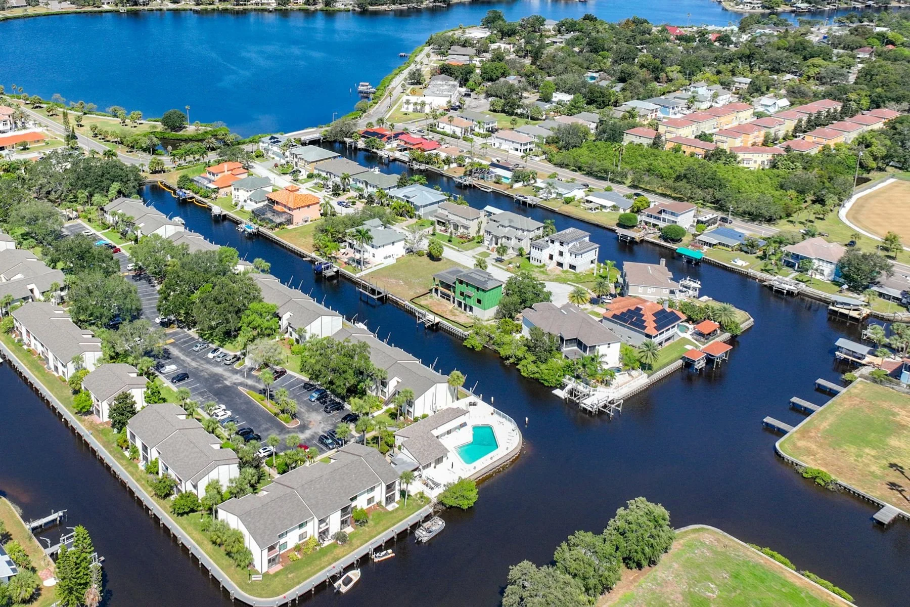 Aerial view of a residential neighborhood with houses along canals, lush greenery, a swimming pool, and a large water body in the background.