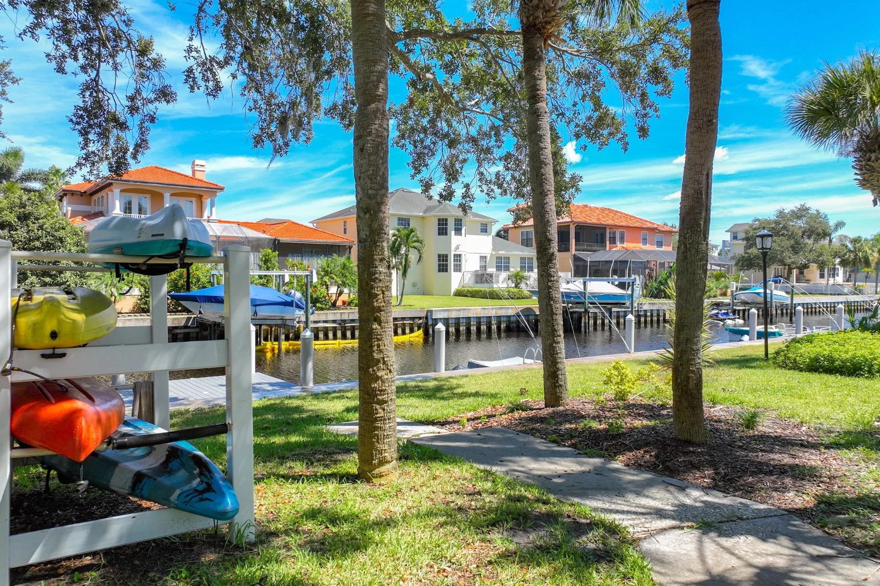 View of a waterfront residential area with colorful houses, boats docked along the waterway, kayaks on a rack, a pathway, trees, and a grassy area under a partly cloudy sky.