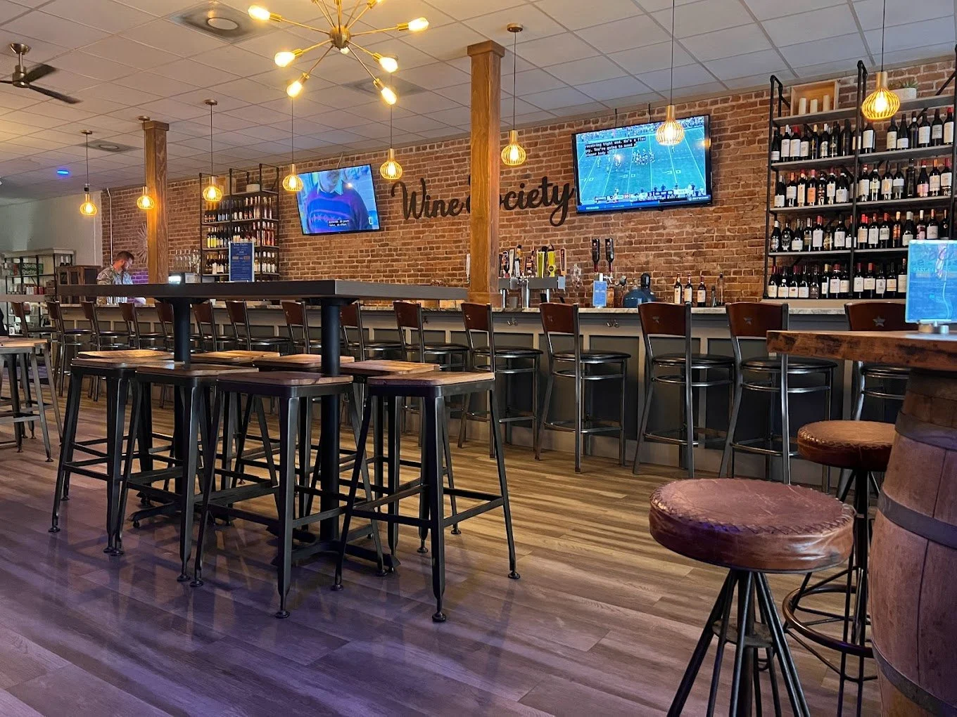 Interior of a wine bar with a wooden bar counter, shelves stocked with wine bottles, high stools, and hanging pendant lights, TV screens displaying sports, and a rustic brick wall with 'Wine Society' sign.