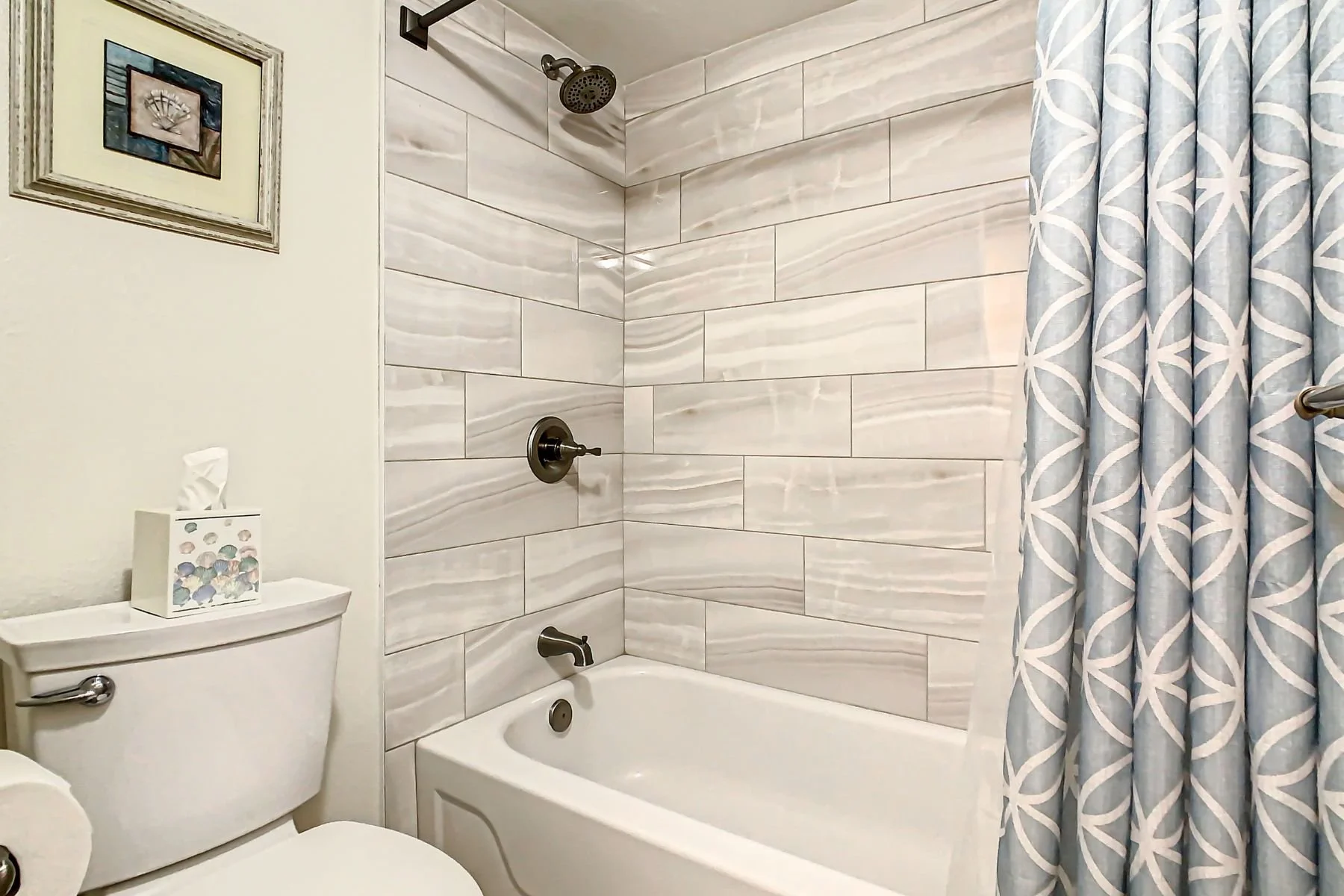 Bathroom with a bathtub, showerhead, and a curtain, with a toilet and tissue box nearby; wall tiles have a marble-like pattern.