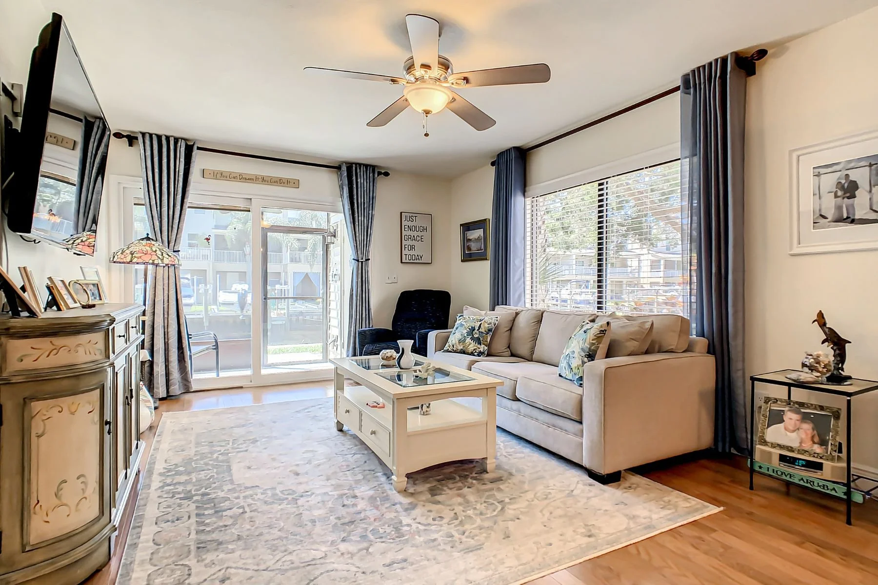 Living room with beige sofa, black armchair, glass-top coffee table, large windows with blue curtains, sliding glass door to patio, wall clock, framed pictures, patterned rug, wooden floor, ceiling fan, TV on wall, side table with photo frame and decorations.