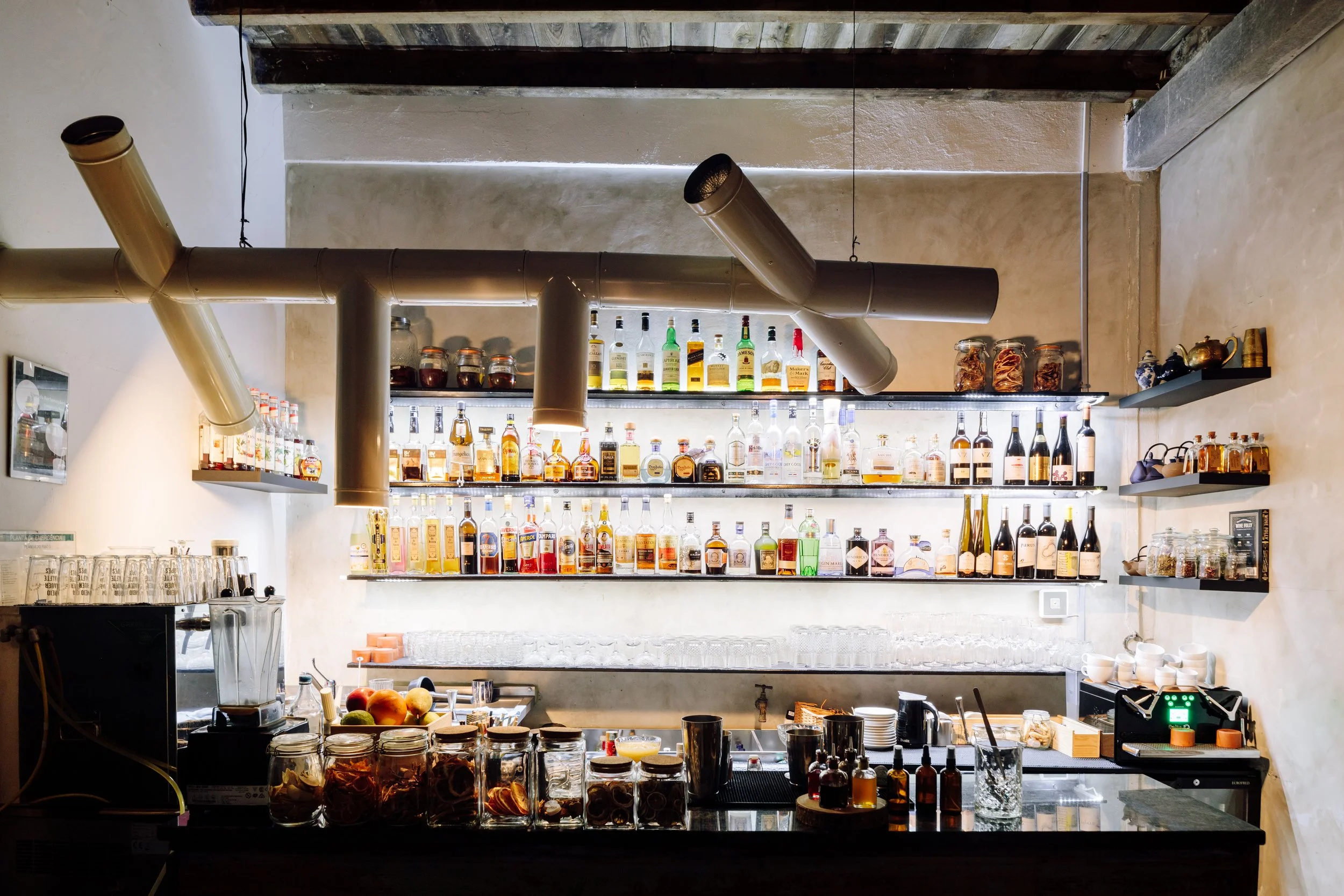 Bar with shelves of various liquor bottles, jars of ingredients, glasses, and bar tools in a cozy cafe or restaurant setting.