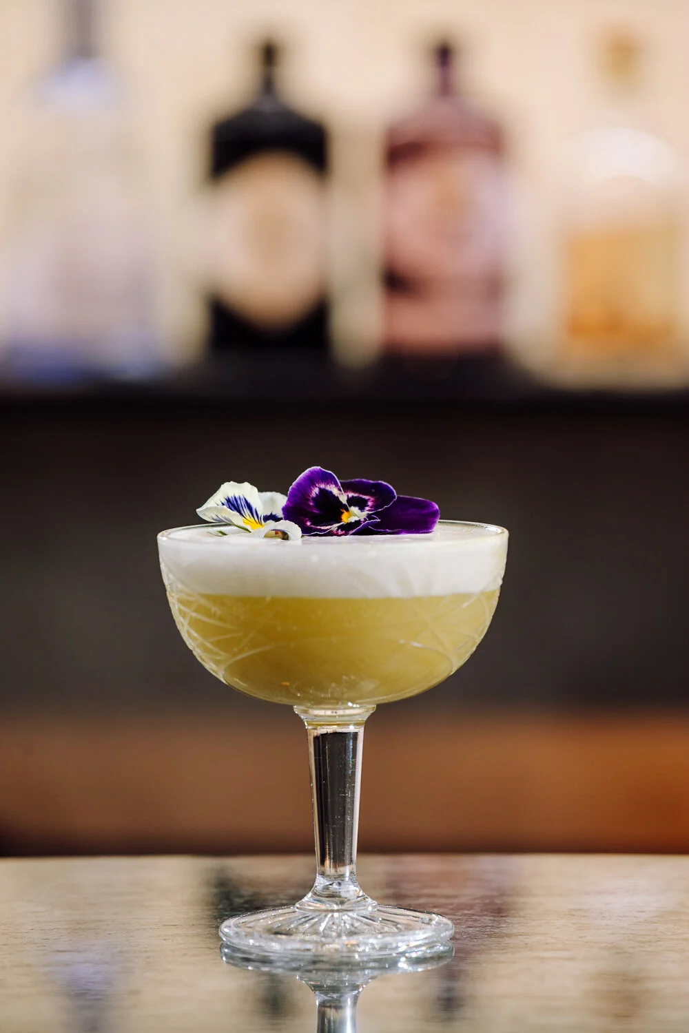 A cocktail in a coupe glass garnished with edible flowers, with blurred bottles in the background.