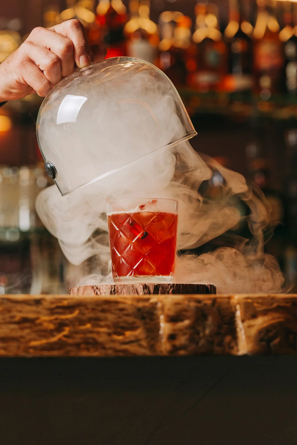 A bartender in a bar pours dry ice into a glass of red cocktail, creating fog and smoke for a dramatic effect.