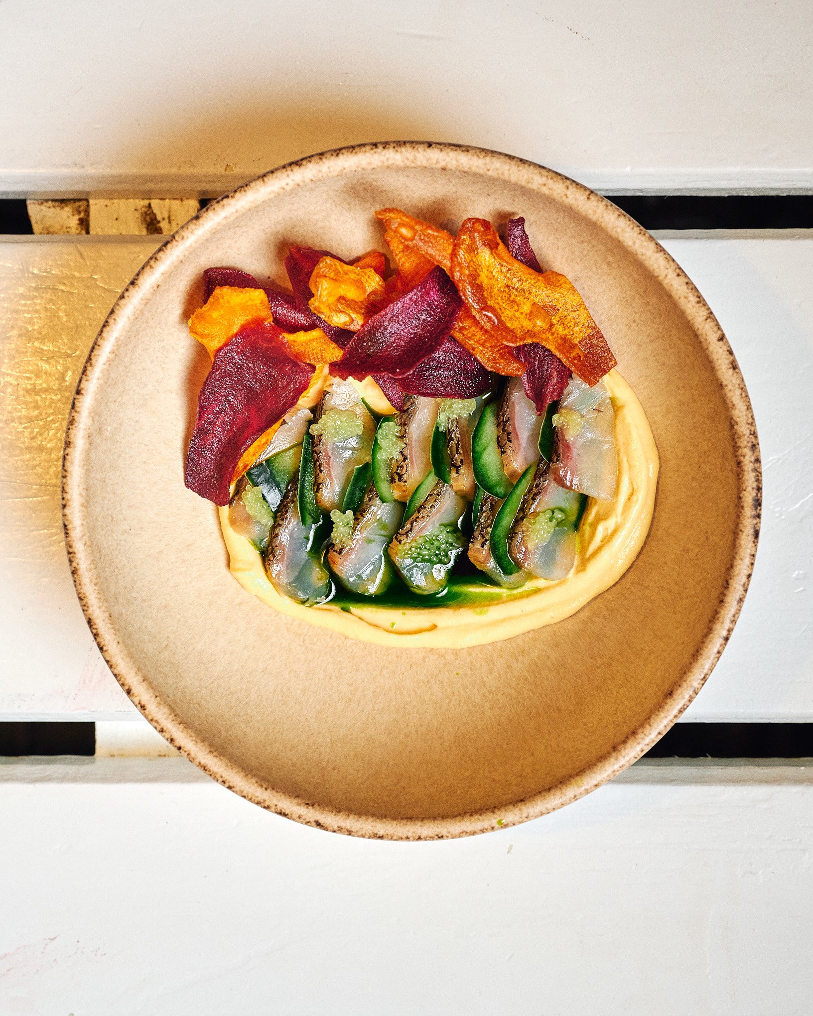 A beige plate with a yellow puree topped with eel slices, green cucumber slices, and crispy bacon strips on the side.