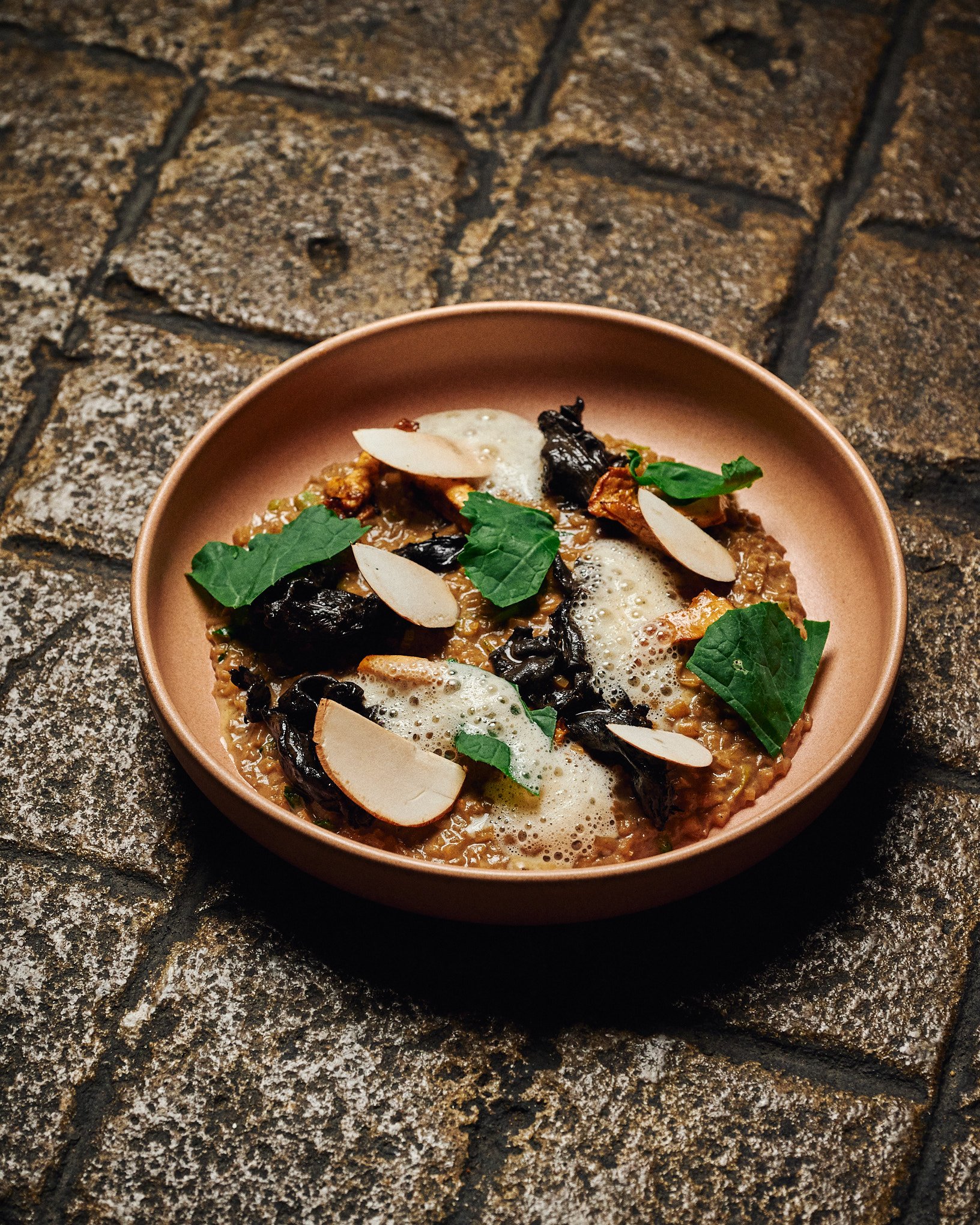 A dish served in a pink bowl containing rice, topped with slices of almonds, fresh green herbs, black wood ear mushrooms, and a foamy sauce.