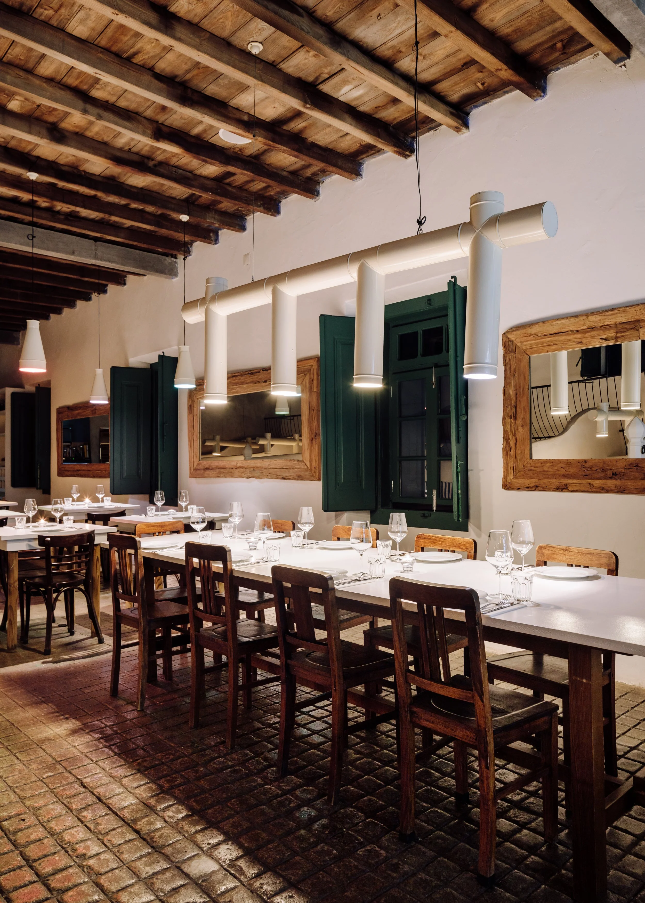 A restaurant interior with a long white table set with wine glasses, plates, and utensils. There are wooden chairs around the table, wooden-framed mirrors on the wall, green window shutters, and a rustic brick floor. The ceiling has exposed wooden be