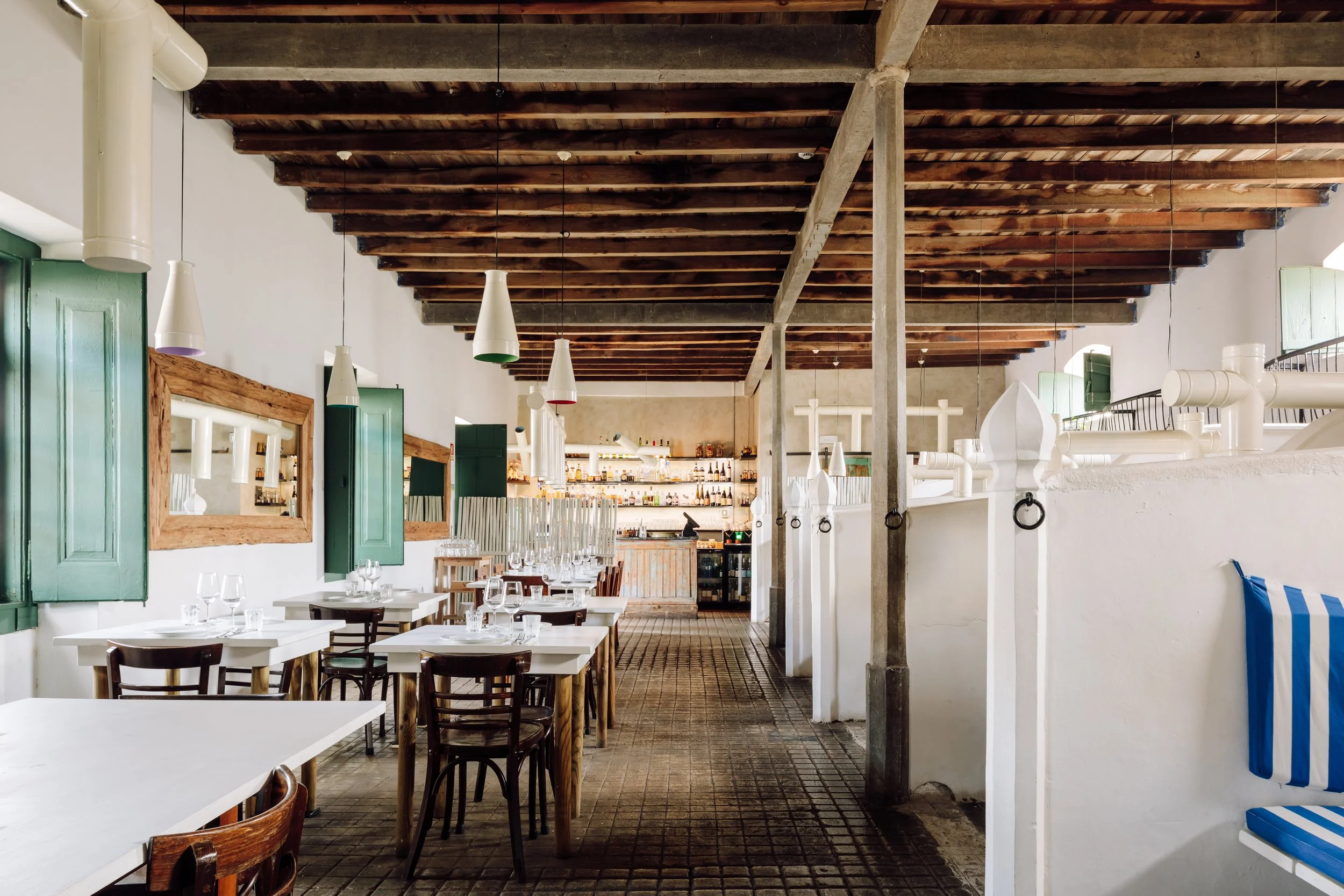 Interior of a rustic restaurant with white walls, green window shutters, wooden ceiling beams, white tables, and dark wood chairs, with a bar in the background.
