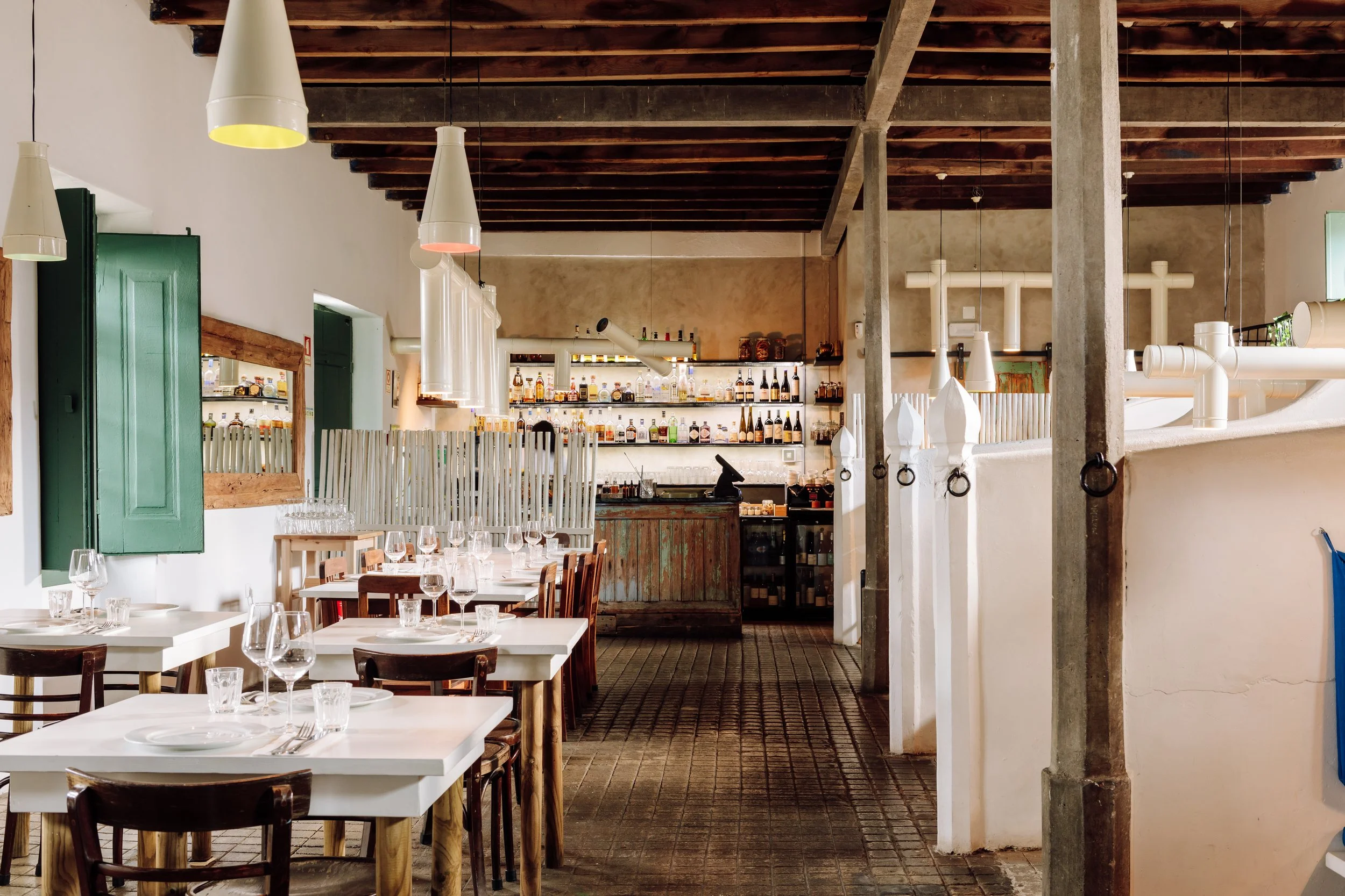 Interior of a rustic restaurant with white tables set with wine glasses, wooden chairs, a bar in the background with bottles on shelves, and decorative lighting fixtures hanging from a wooden ceiling.