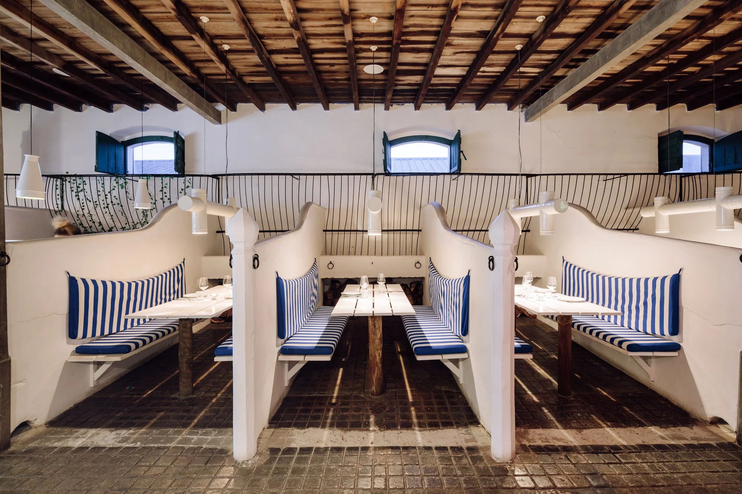 Interior of a restaurant with white booths with blue and white striped cushions, a wooden table in each, and overhead white pendant lights. The background shows a white wall with three small windows, green shutters, and a black metal railing.