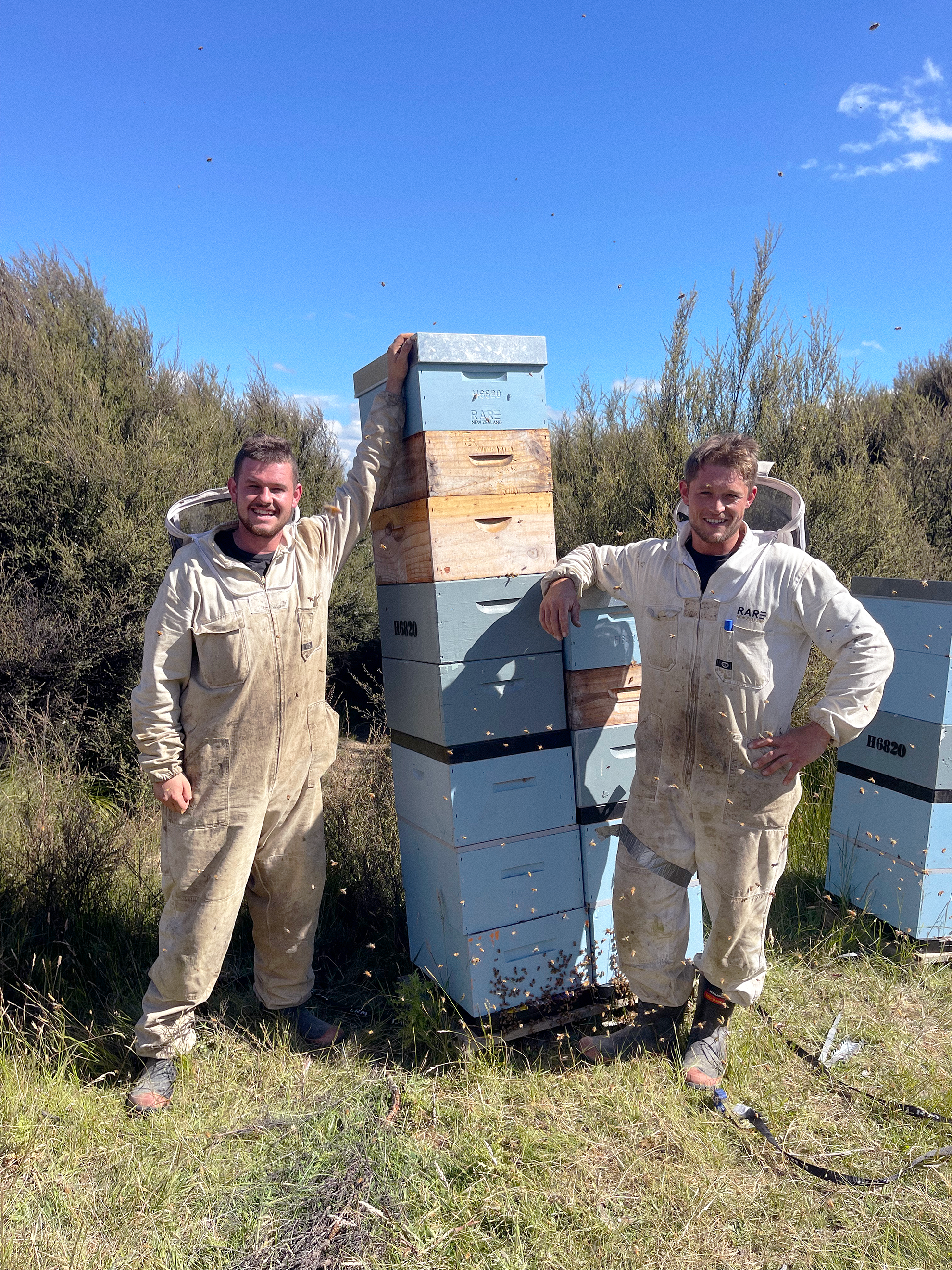 Two men in work suits standing outdoors in front of beehives on a sunny day, surrounded by tall grass and bushes.