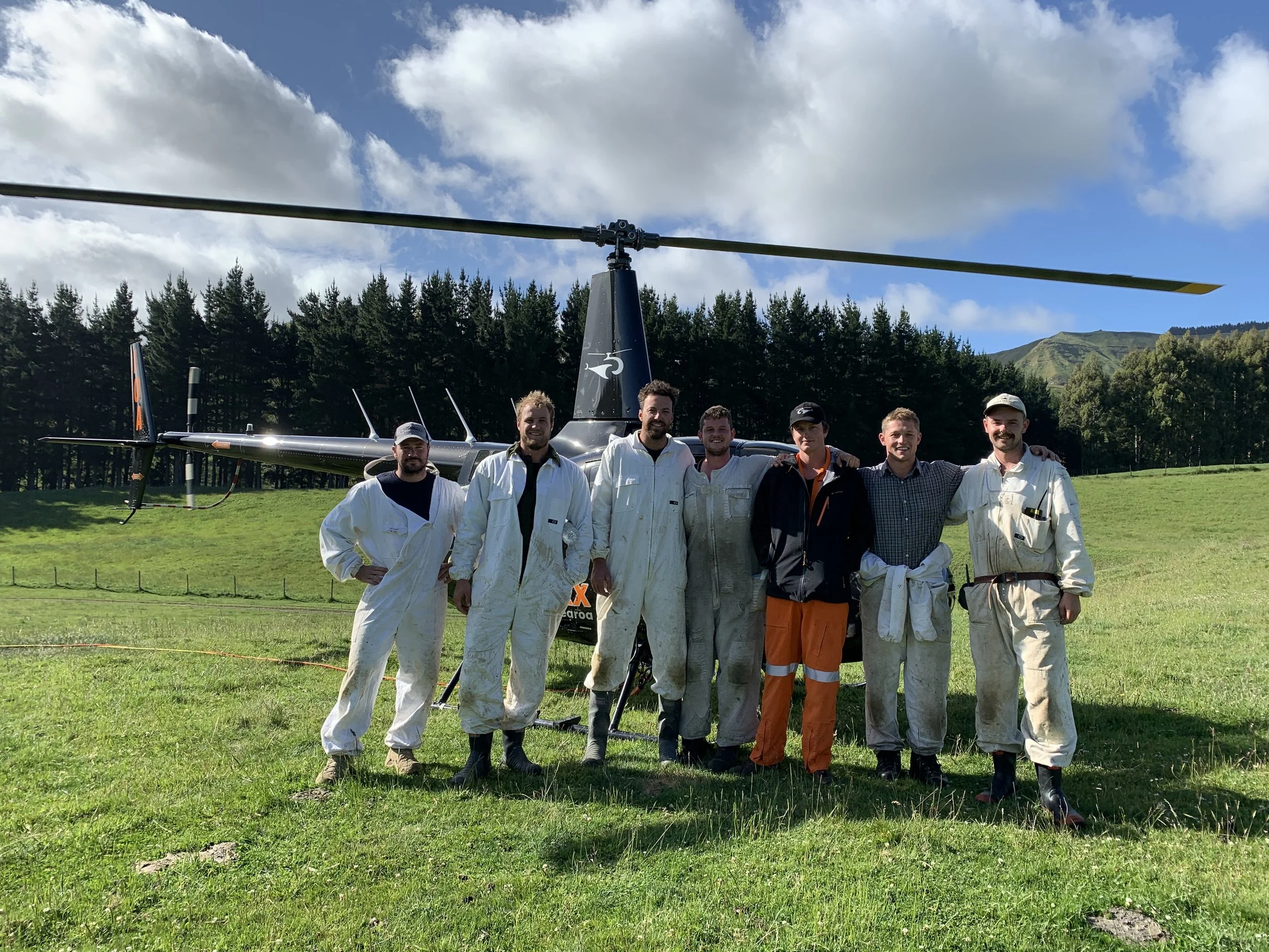 A group of seven men standing outdoors in front of a helicopter, with mountains and a forest in the background, under a partly cloudy sky.