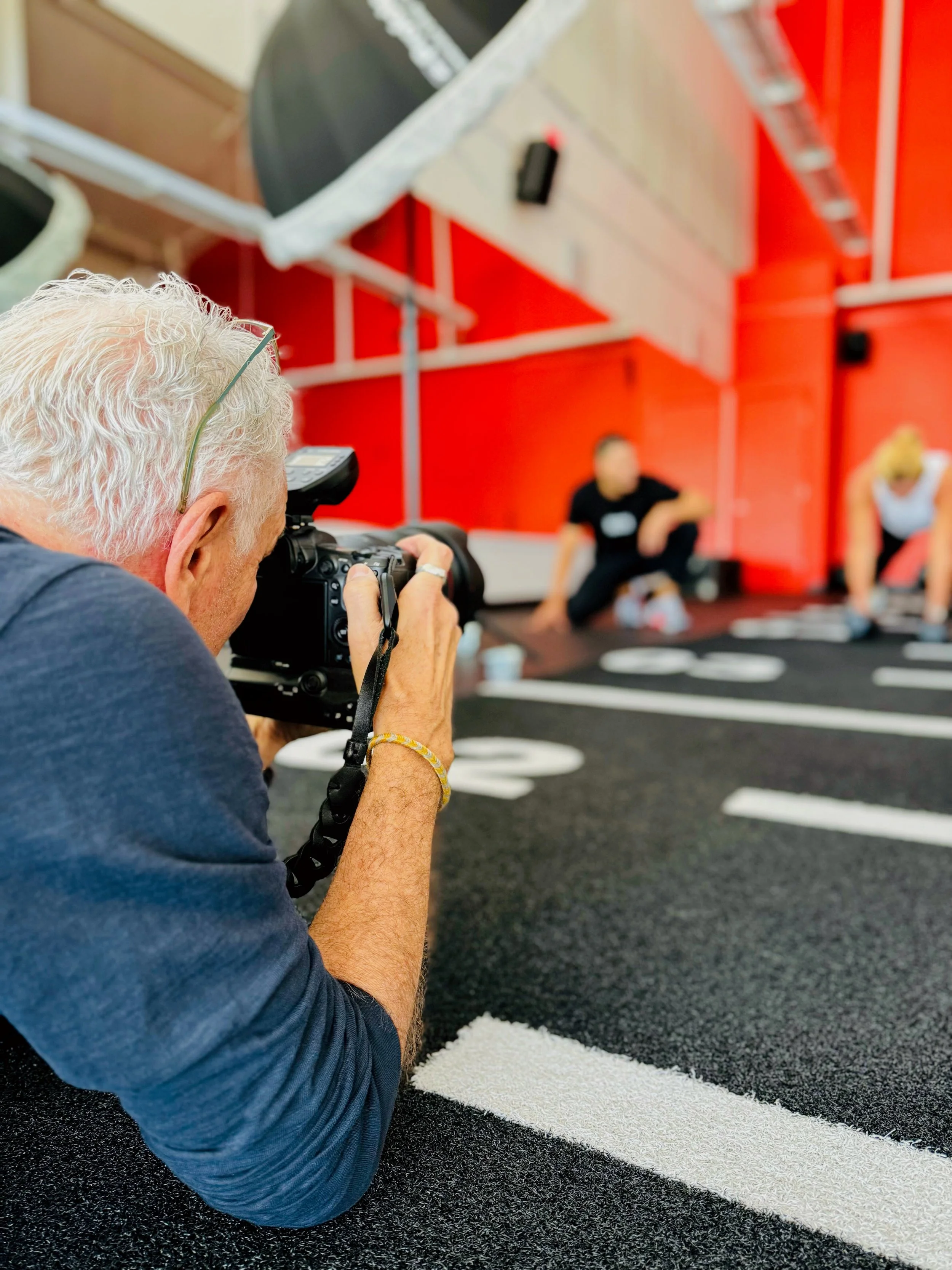 An older man with white hair is taking a photograph of two women doing yoga on a black mat in a gym with red walls.