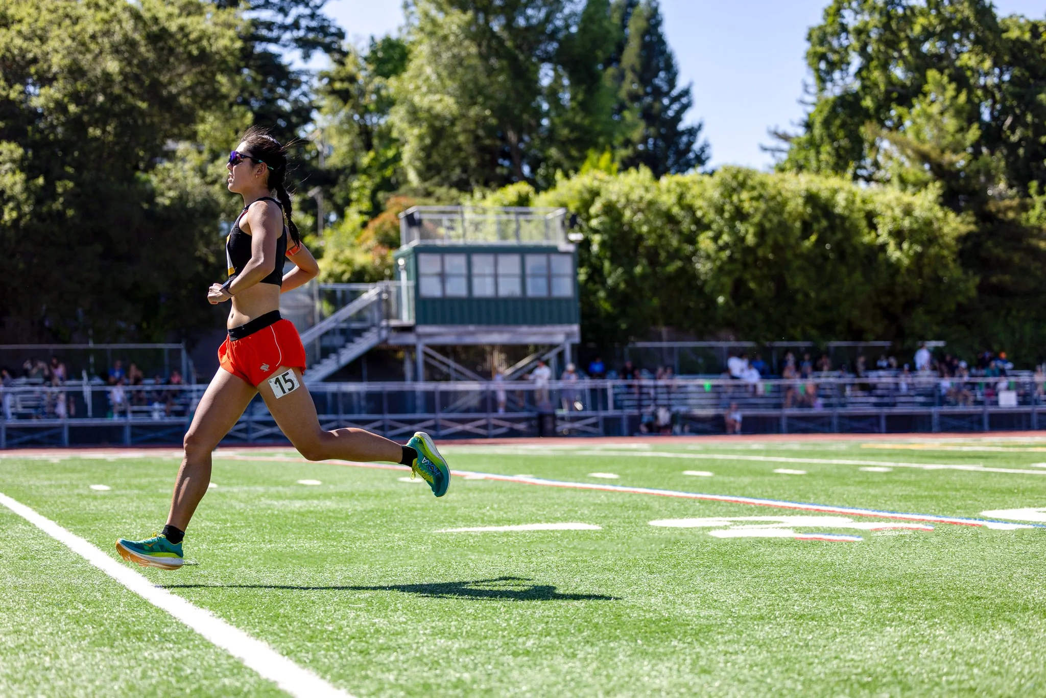 A female athlete running on a green track during a race, wearing red shorts, a black sports top, sunglasses, and running shoes, with spectators watching in the background on a sunny day.