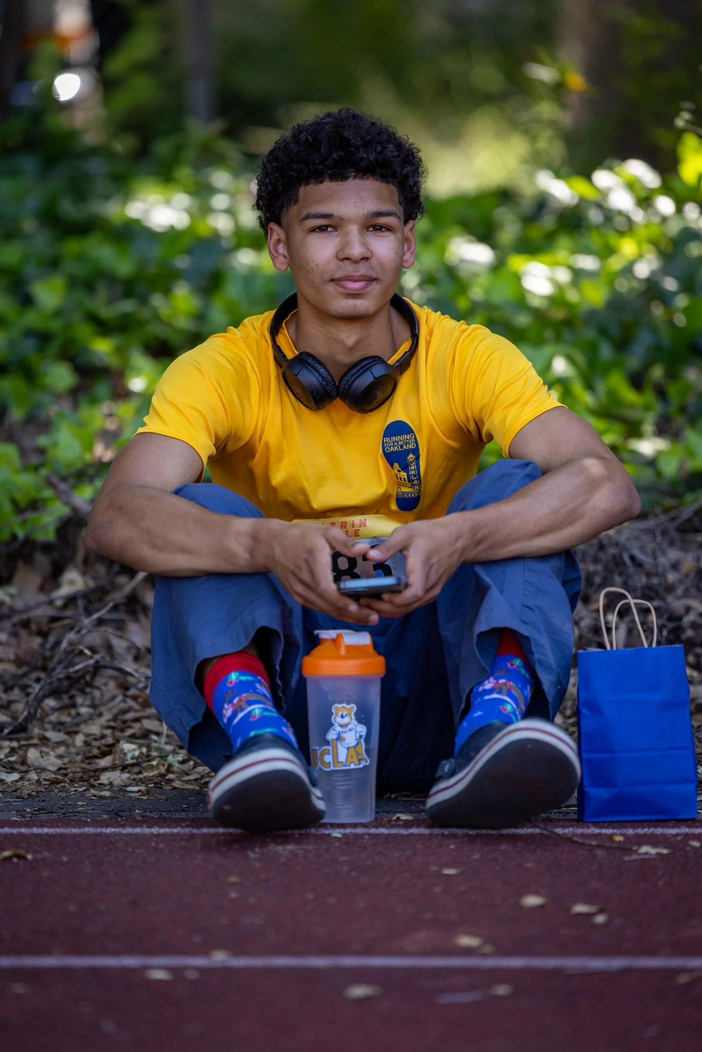 A young man sitting on the ground outdoors, wearing a yellow shirt, blue pants, and colorful socks, with headphones around his neck, holding a smartphone, and sitting next to a water bottle and a blue gift bag.