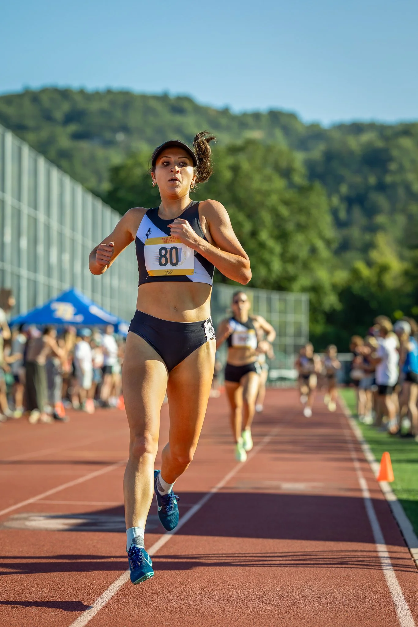 A female athlete running on a track during a race, wearing a black sports bra, black shorts, and a black visor helmet, with the number 80 on her bib, and surrounded by spectators and other runners.