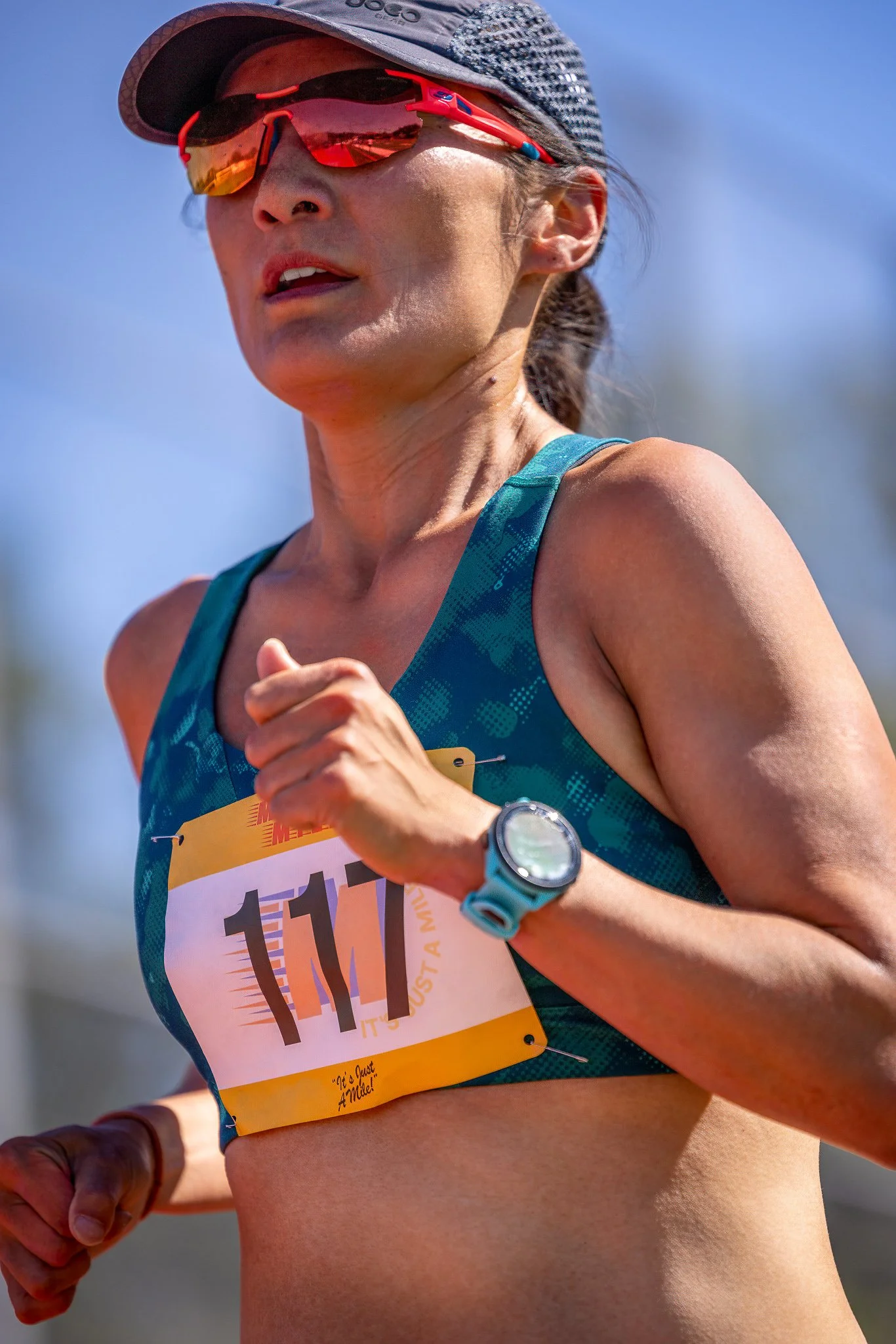 Close-up of a female runner with a race bib number 111, wearing sunglasses, a cap, a sports watch, and a teal workout top, running outdoors under clear blue sky.