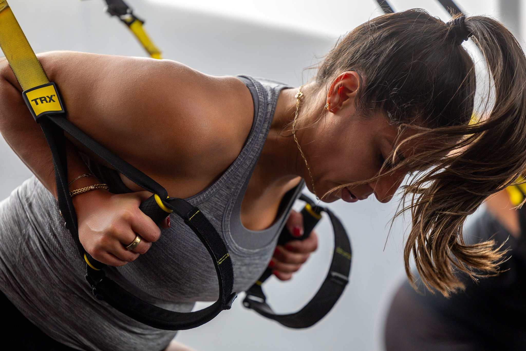 Woman with brown hair in a ponytail, wearing a gray tank top, doing an intense workout with TRX suspension straps at a gym.