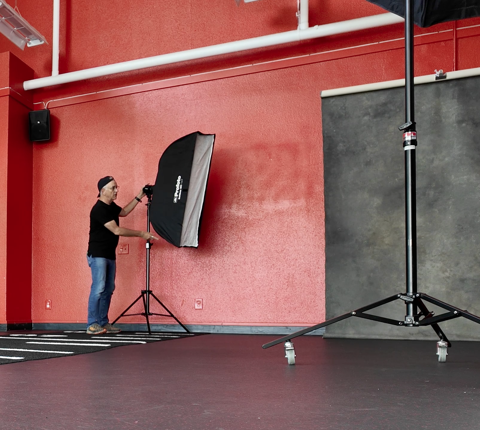 A photographer wearing a black shirt, blue jeans, gray shoes, and a backwards cap is setting up lighting equipment in a studio with a red textured wall, black and white striped carpet, and grey backdrop.