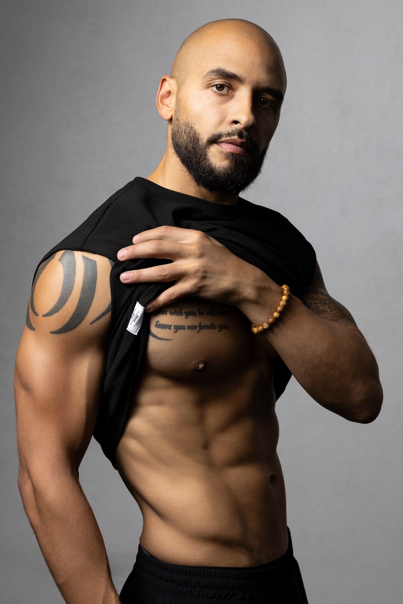 A muscular bald man with a beard poses with his shirt lifted, showing tattoos on his chest and arm, against a gray background.
