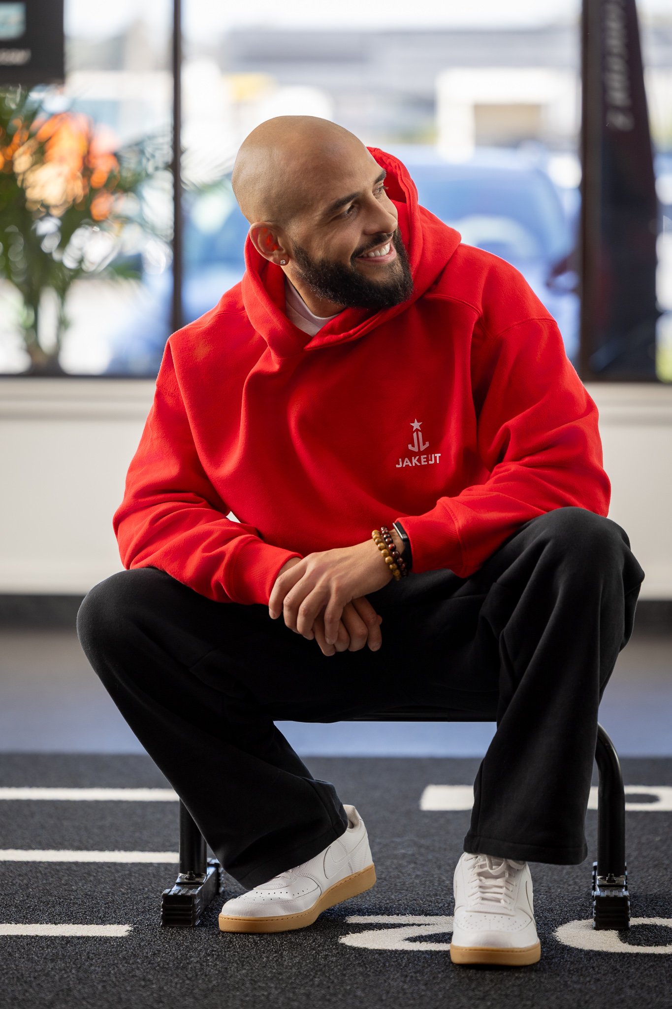 A bald man with a beard sits on a small black stool inside a gym or fitness center, wearing a red hoodie with the logo 'JAKET' and a star, black pants, and white sneakers. He is smiling and looking to his right with a cheerful expression.