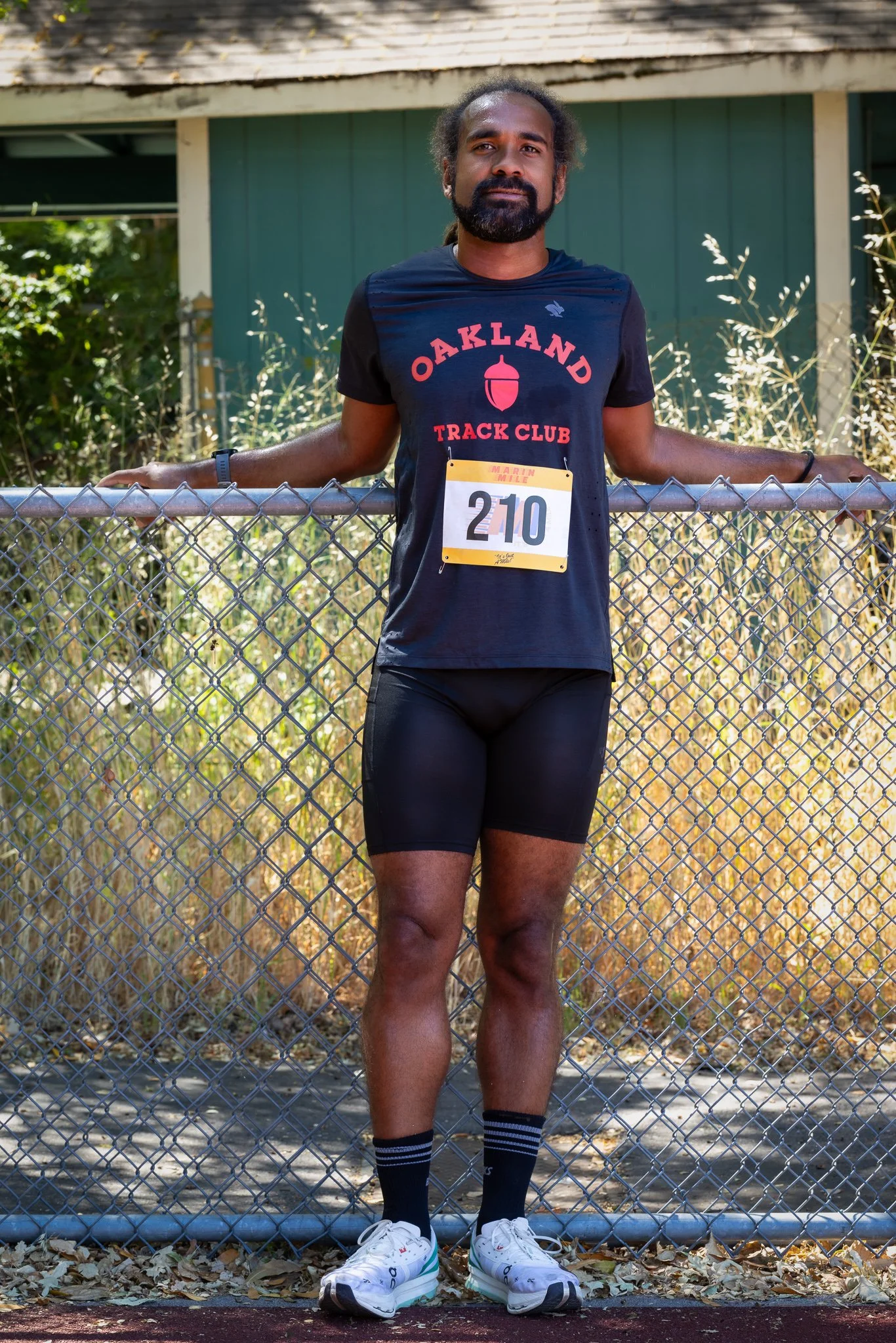 A man standing behind a chain-link fence wearing a black Oakland Track Club t-shirt, black shorts, black socks with white and blue stripes, and white running shoes, with a race bib number 210 attached to his shirt.