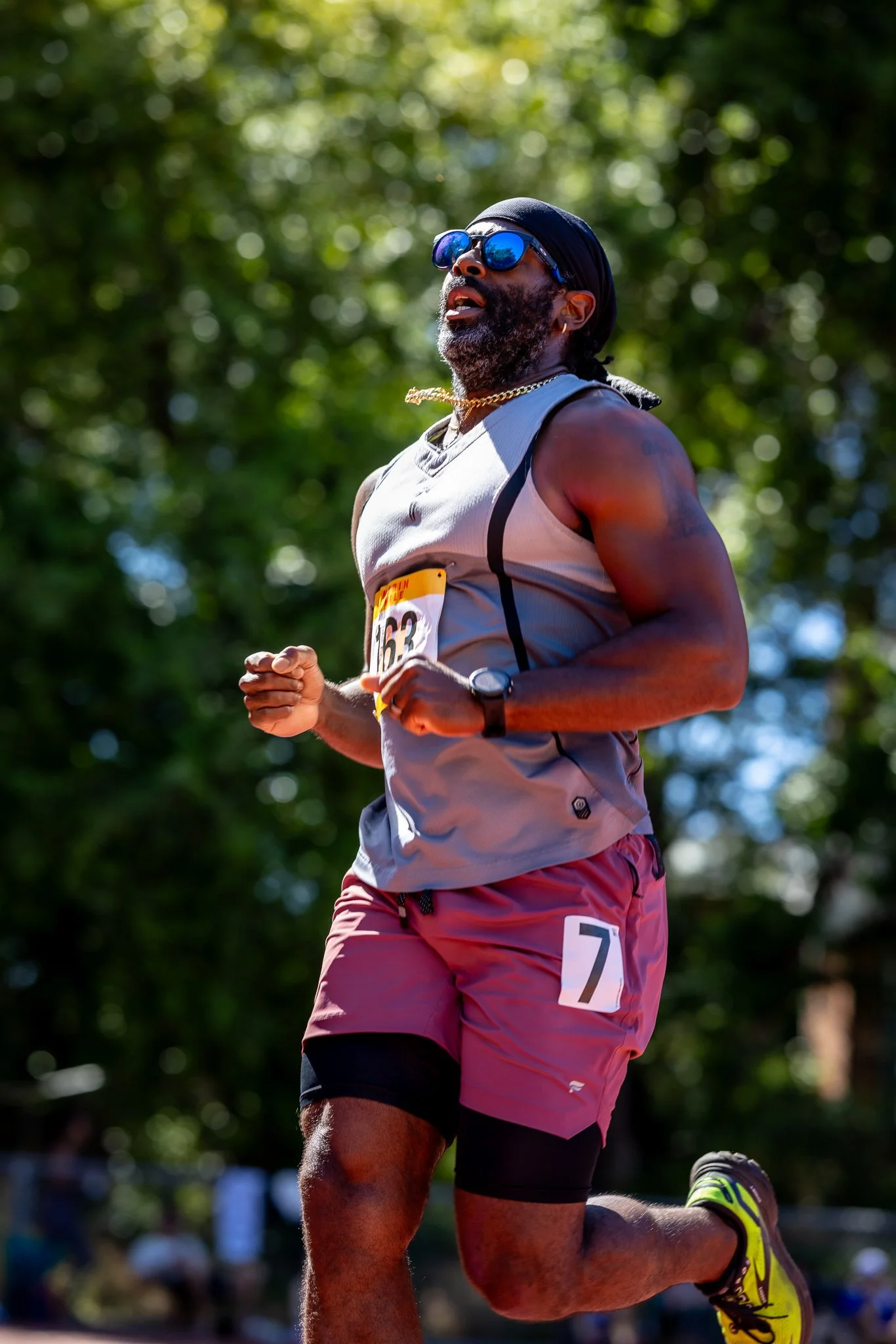 A man in athletic gear running outdoors during a race, wearing sunglasses, a headband, a bib number 63, and a watch, with a background of green trees.
