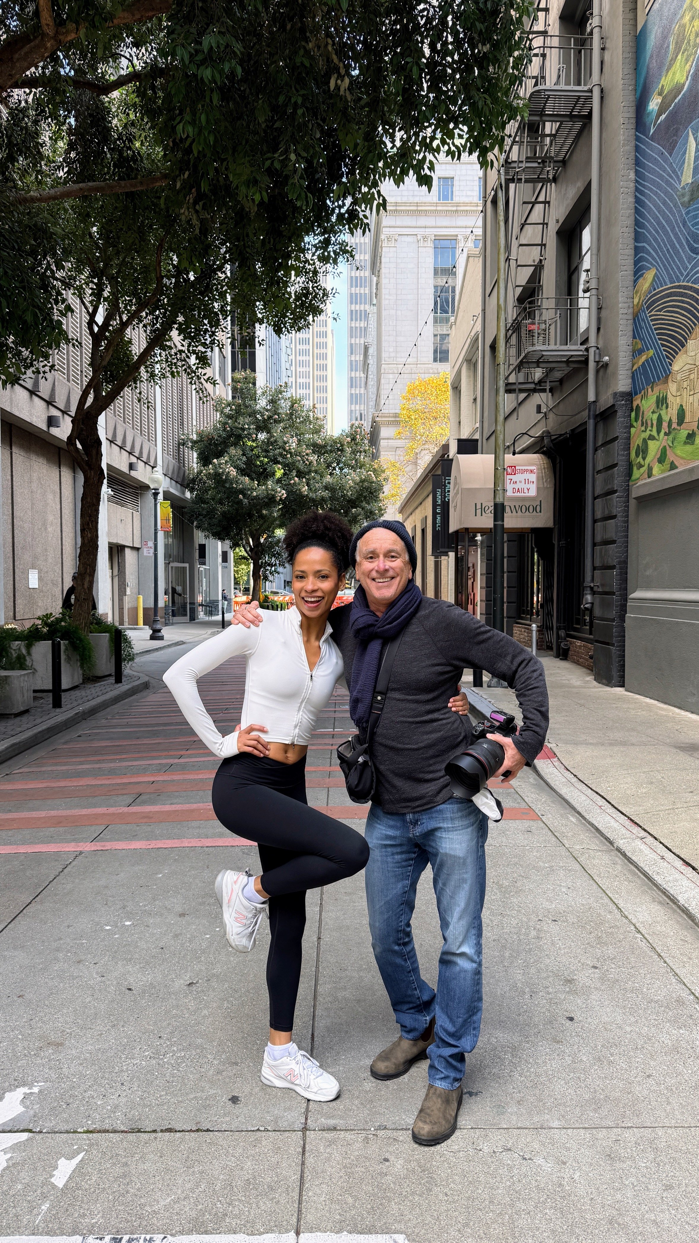 A woman and a man stand together on a city sidewalk, smiling at the camera. The woman has dark curly hair in a puff, is wearing a white zip-up jacket and black leggings, and has her arm around the man. The man, wearing a black beanie, gray sweater, a