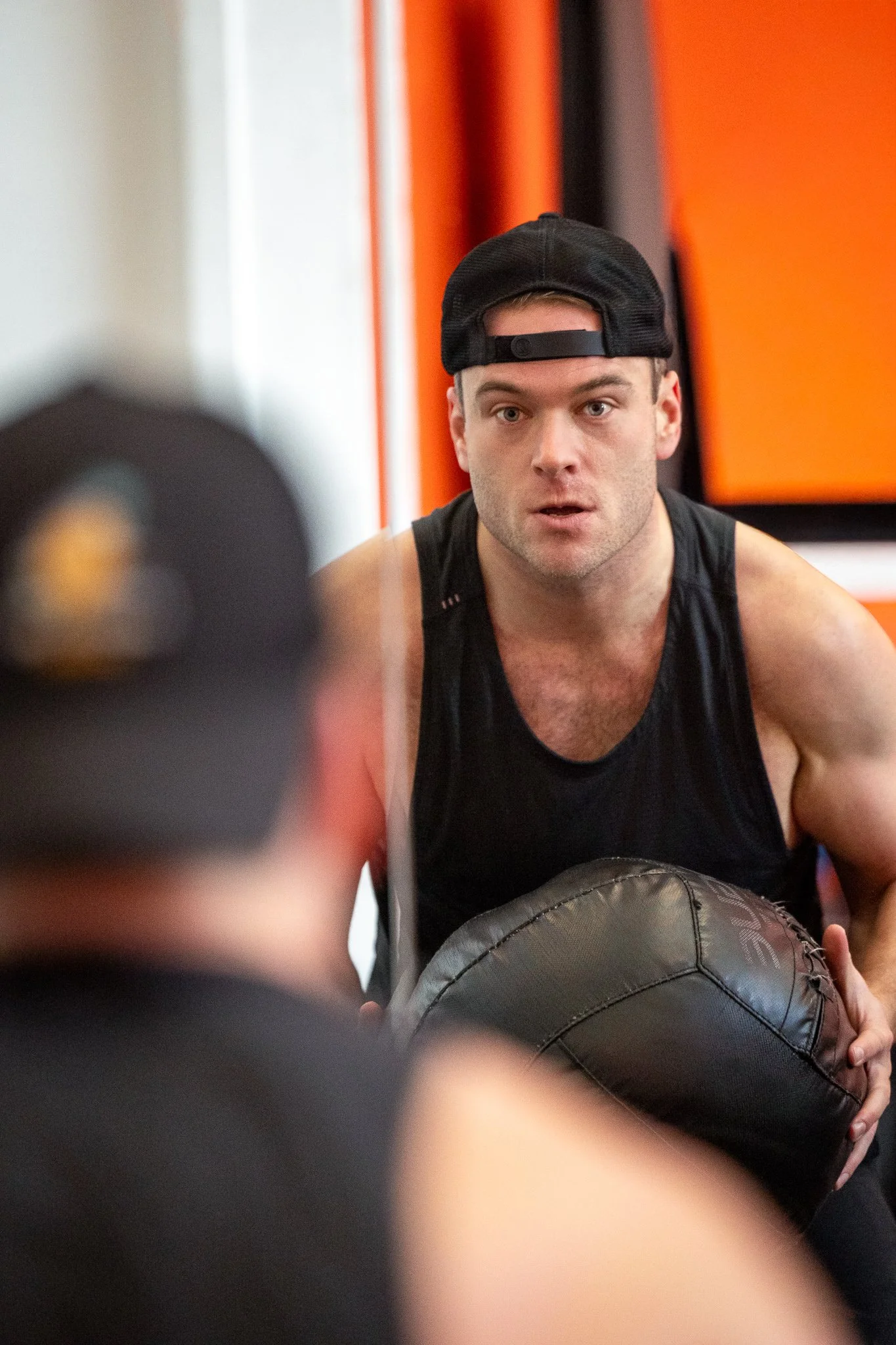 Man in black sleeveless shirt and black baseball cap working out with a medicine ball in a gym.
