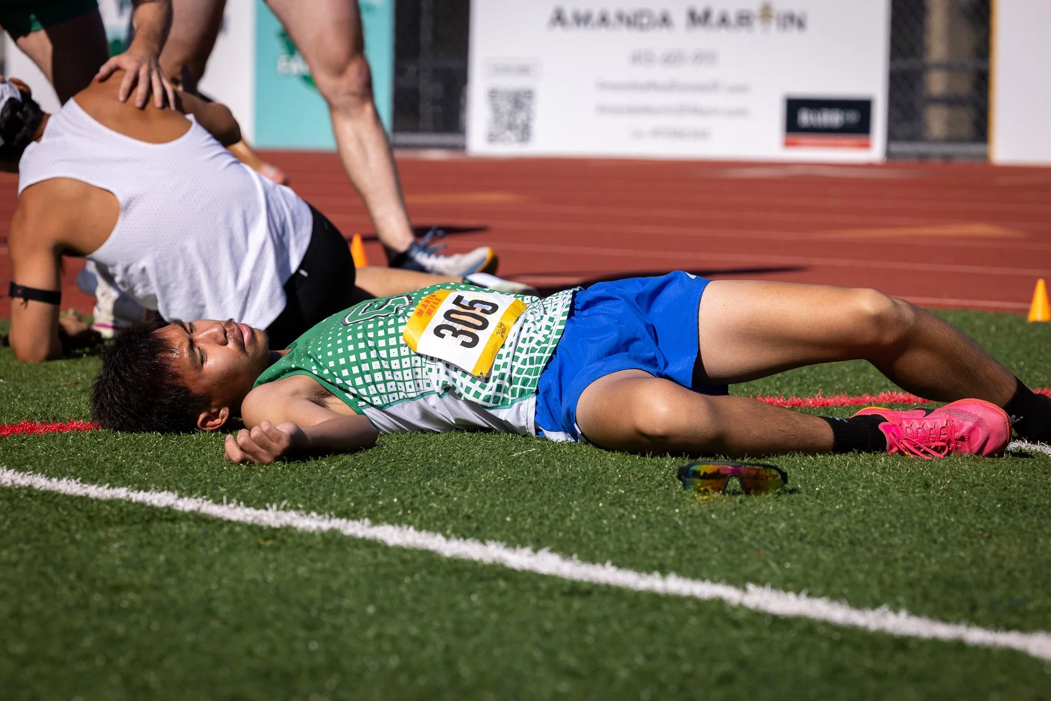 A male track athlete wearing a green and white uniform with the bib number 3055 lies on the ground, appearing to be exhausted or in pain, beside other athletes stretching or resting after a race on a track field.