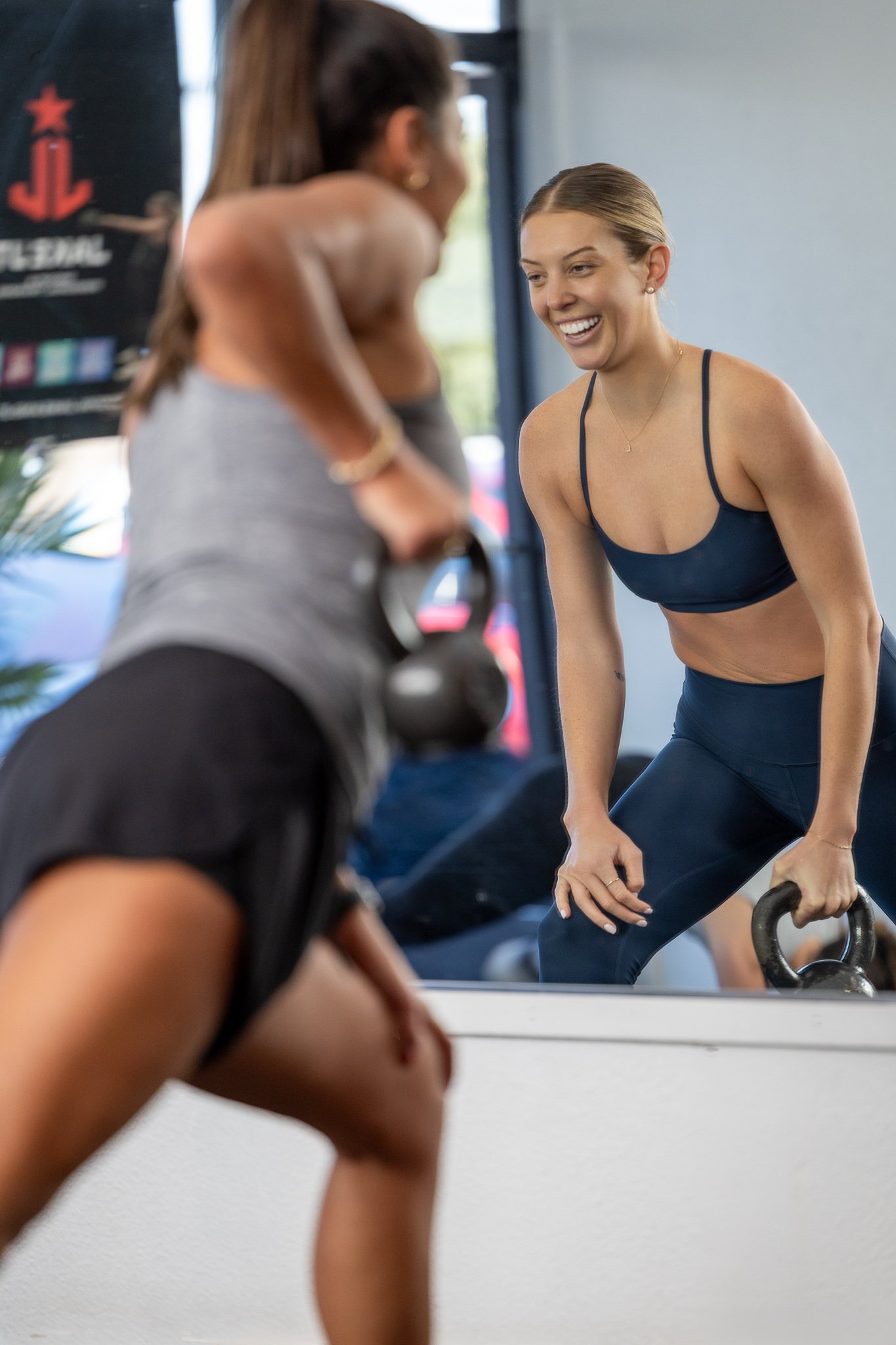 Two women working out with kettlebells at a gym, one in a gray tank top and black shorts, and the other in a navy sports bra and matching leggings, smiling at each other