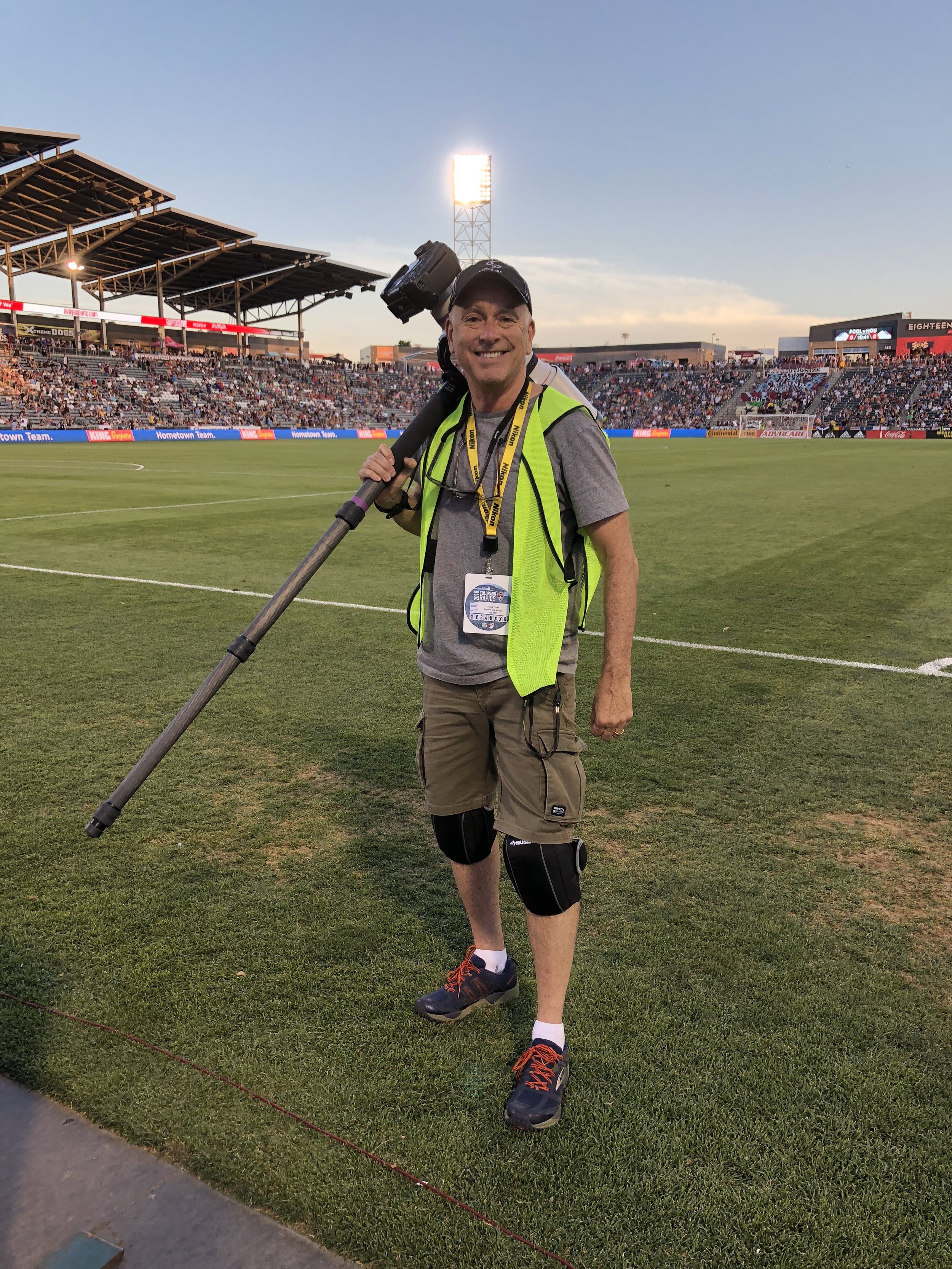A man standing on a soccer field during a match, carrying a camera on his shoulder, wearing a bright vest, shorts, knee braces, and sneakers, with a large crowd in the stadium behind him.